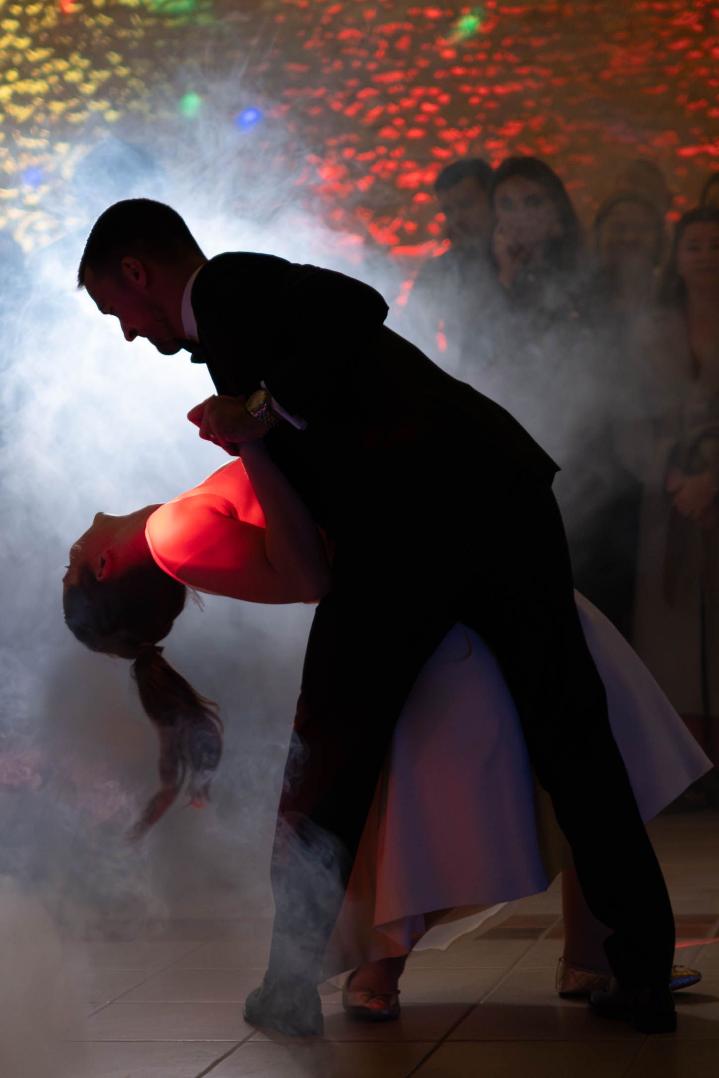Silhouetted dancers perform dramatic dips and spins on a foggy dance floor with red and yellow disco lights overhead.