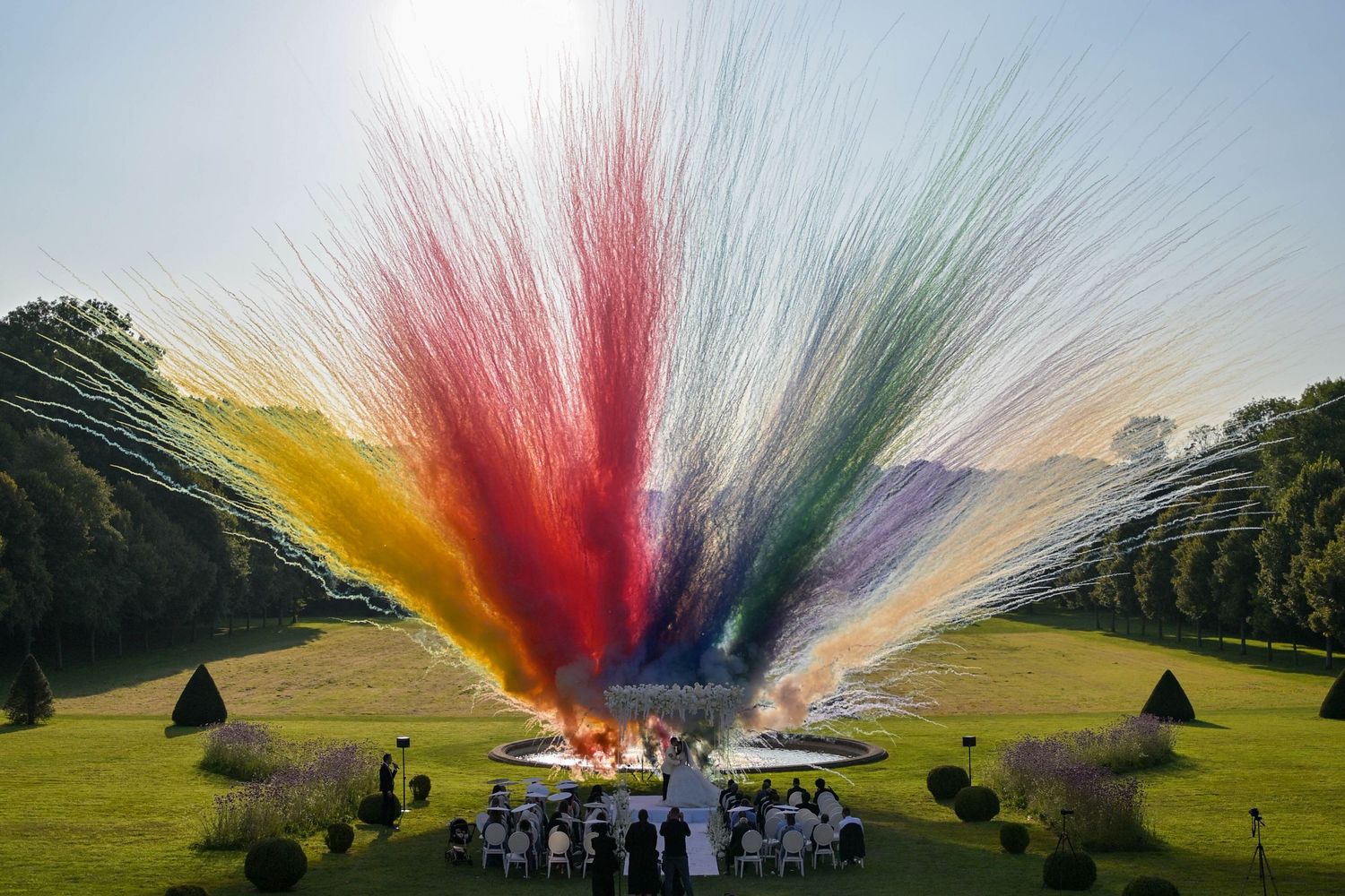 A sequence of colorful powder explosions against a green hillside landscape creates a vibrant rainbow effect in the sky.