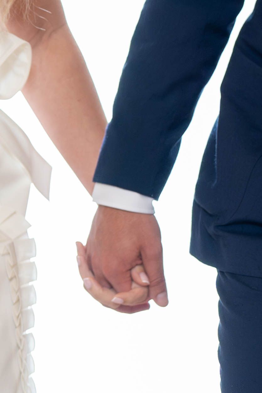 Two people wearing formal attire hold hands in front of a white background.