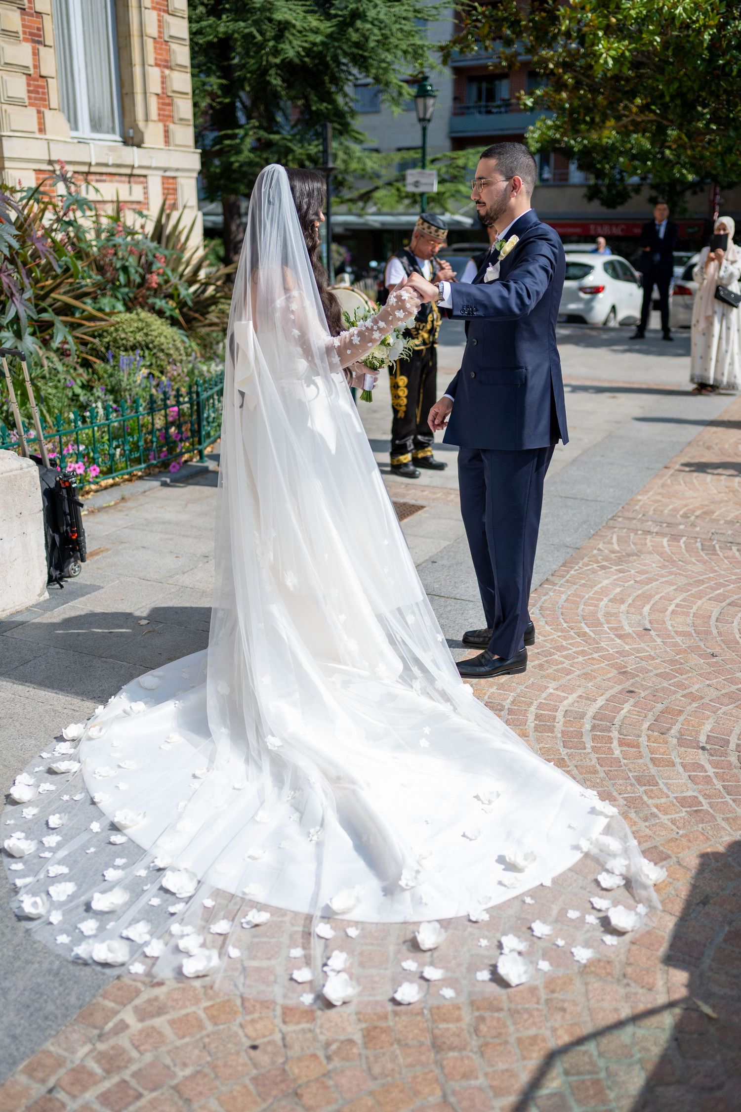 A bride in a flowing white dress with a long veil stands on brick pavement while being photographed on her wedding day.