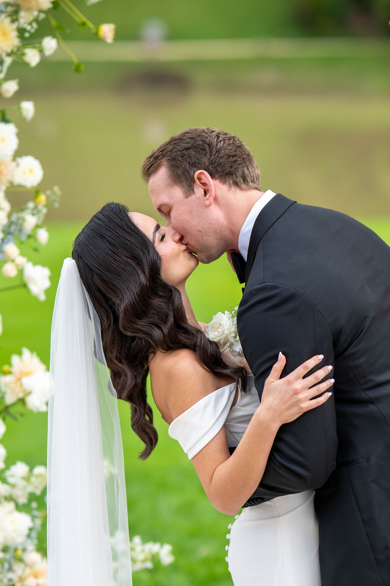 A romantic wedding kiss between newlyweds surrounded by white flowers in a garden setting.