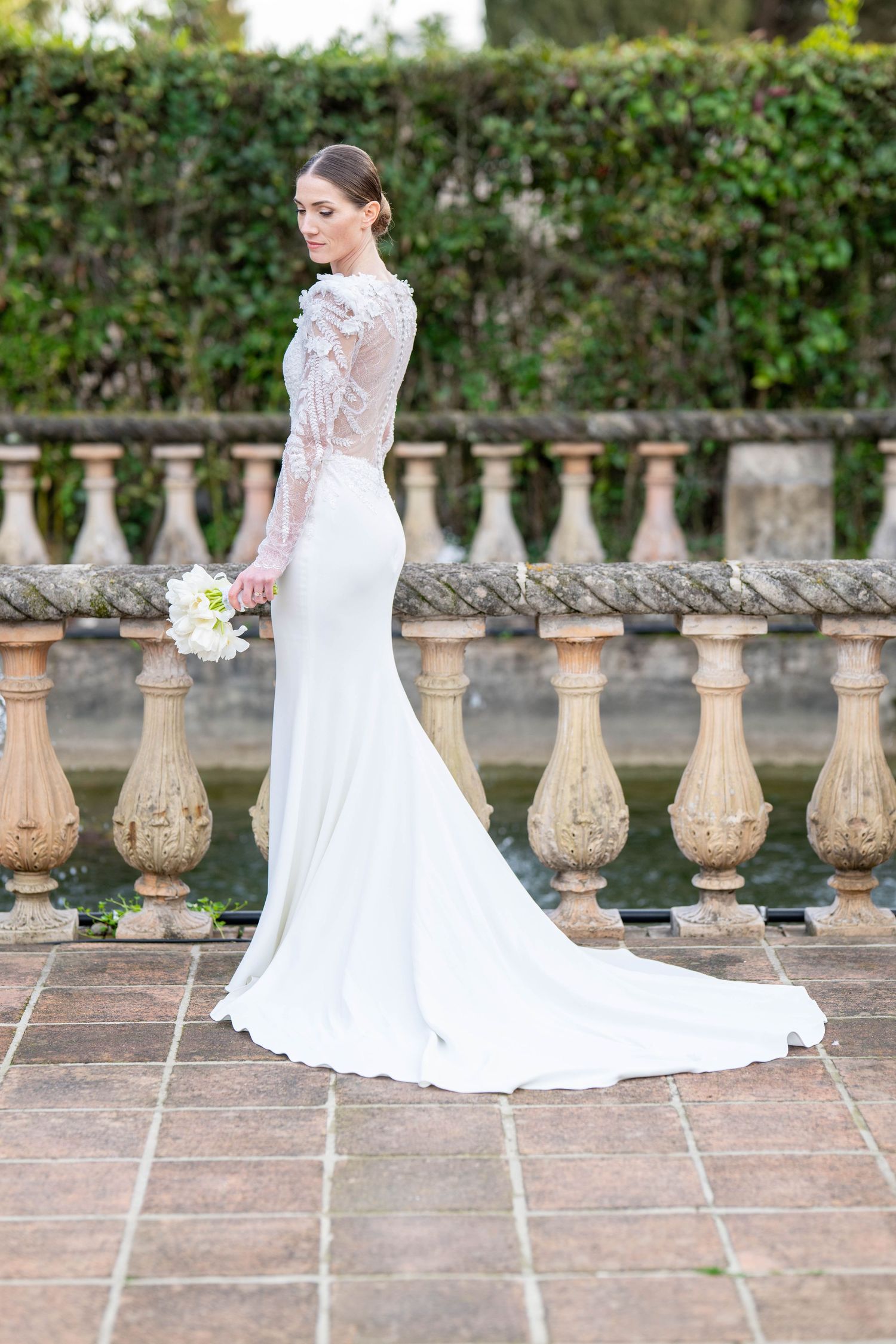 Bride in lace wedding gown with flowing train poses on stone terrace.