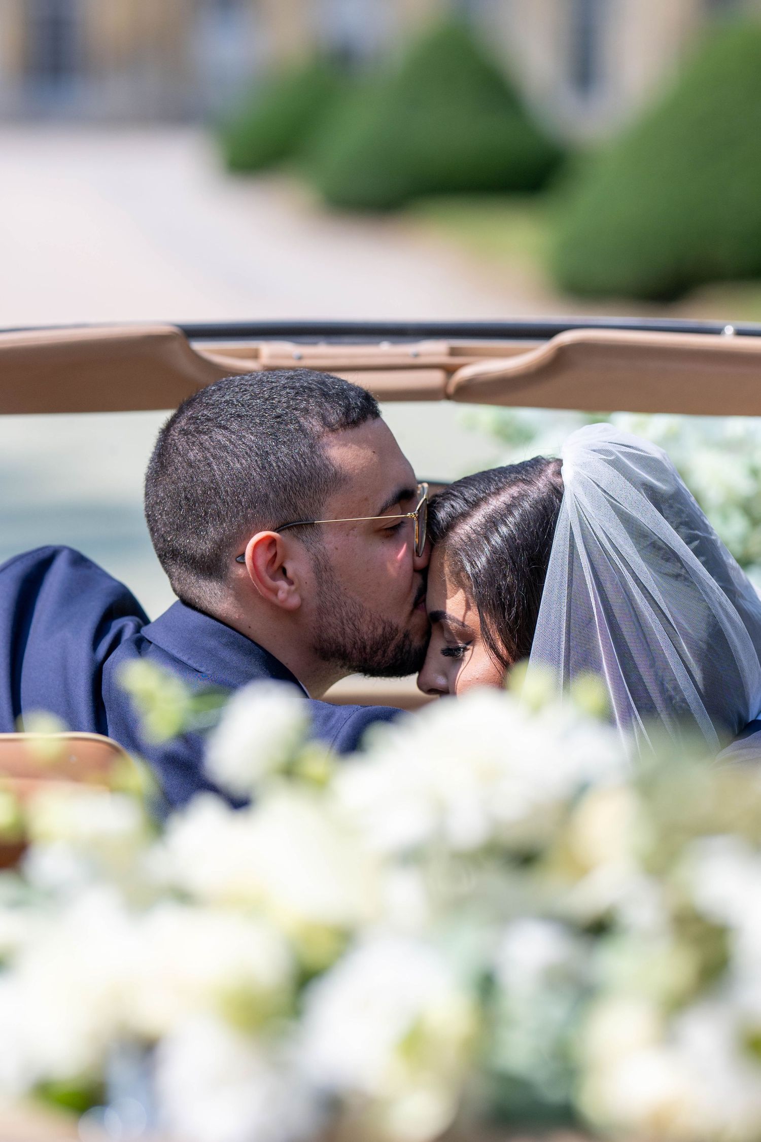 A couple shares an intimate kiss outdoors with white flowers and greenery in the background.