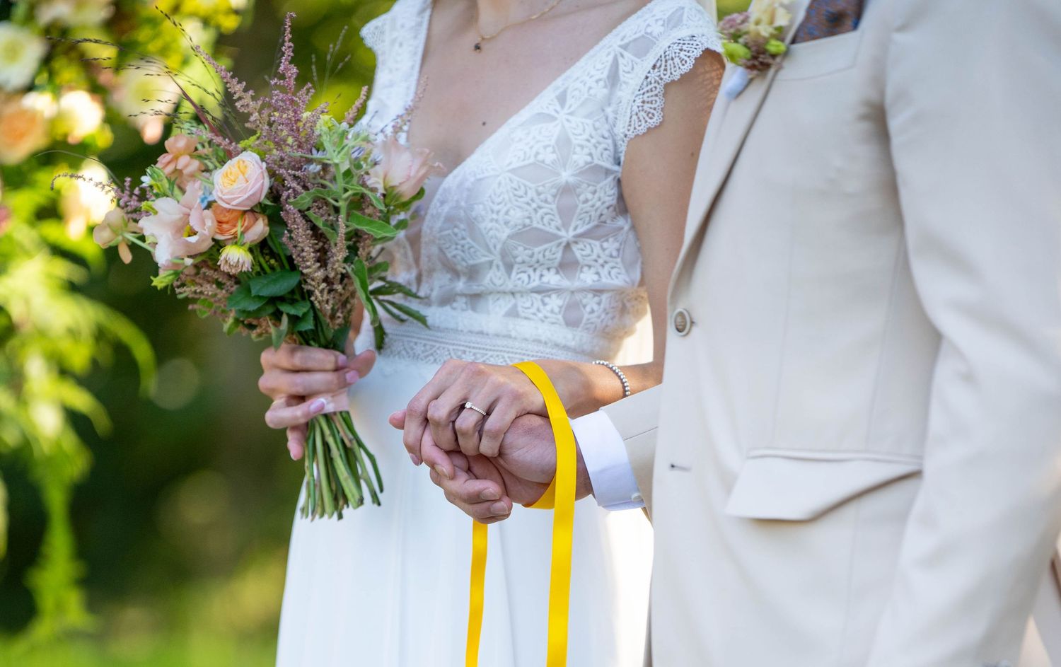 Elegant wedding couple holds yellow ribbons while walking through a lush garden setting.