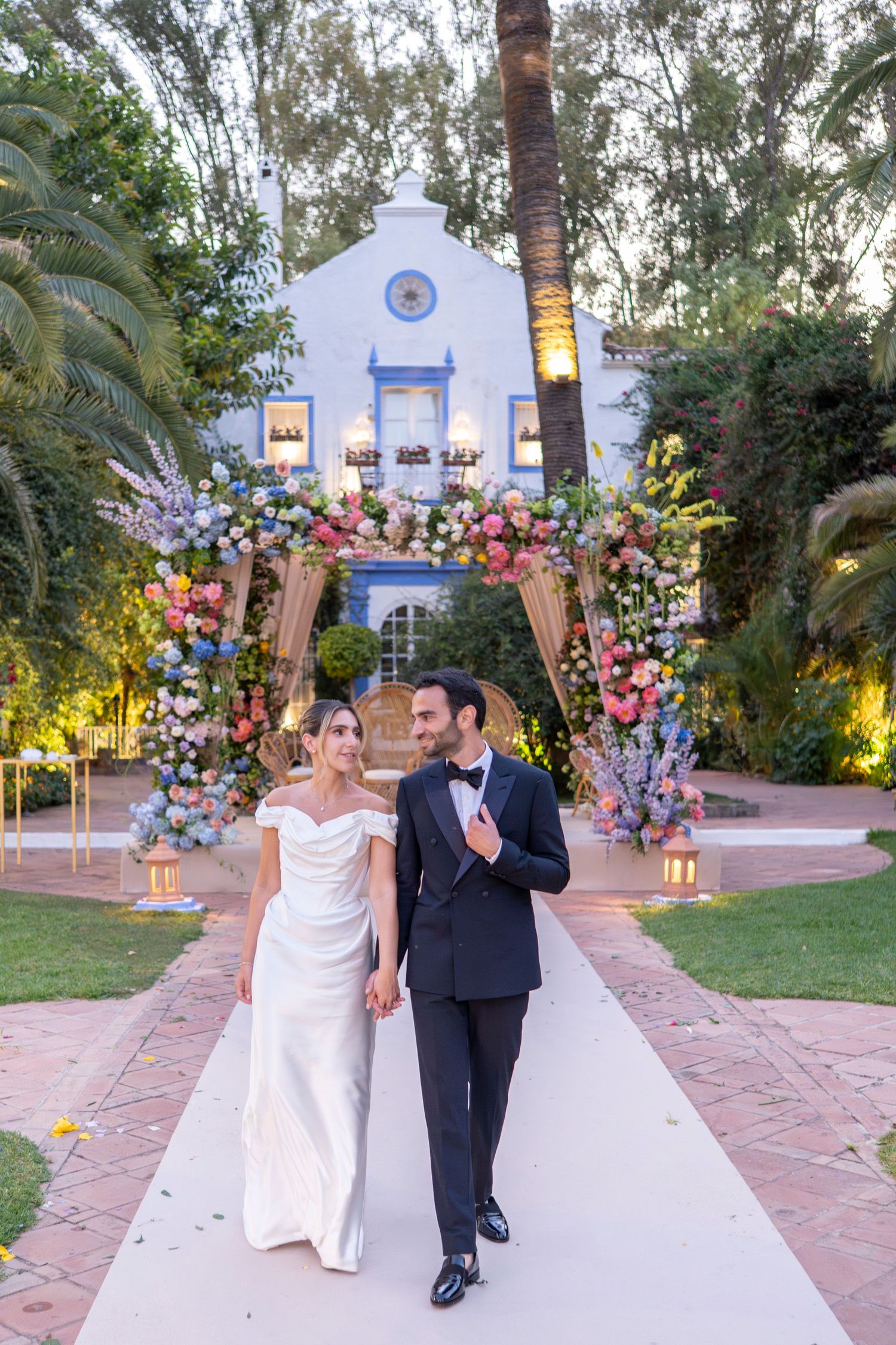 A couple walks down a white pathway at a Spanish-style venue decorated with colorful floral arrangements and lanterns at dusk.