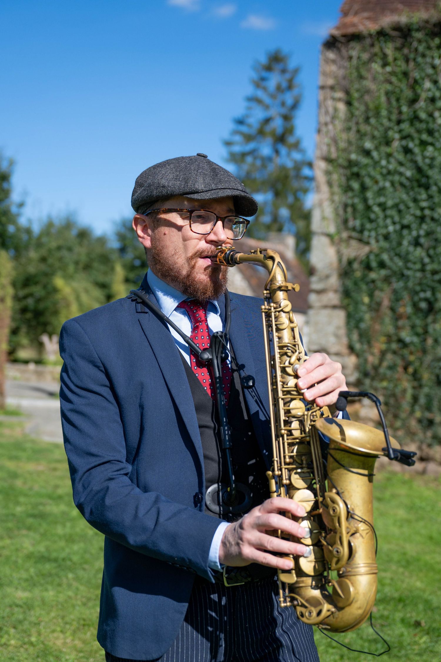 A saxophone musician in a navy suit and red tie performs outdoors on a sunny day in a grassy field.
