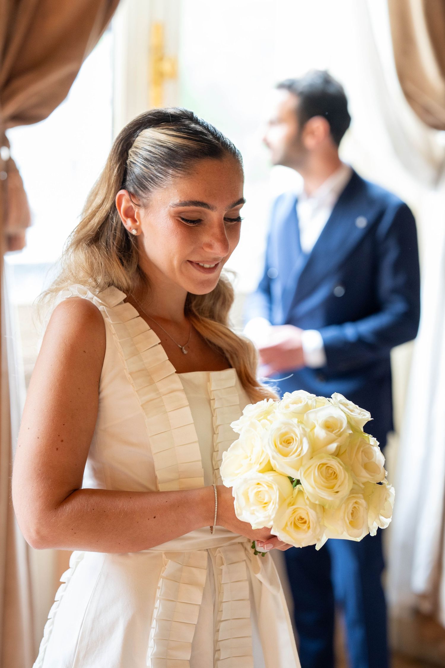 Bridesmaids in white dresses hold elegant bouquets of cream-colored roses at an indoor wedding celebration.