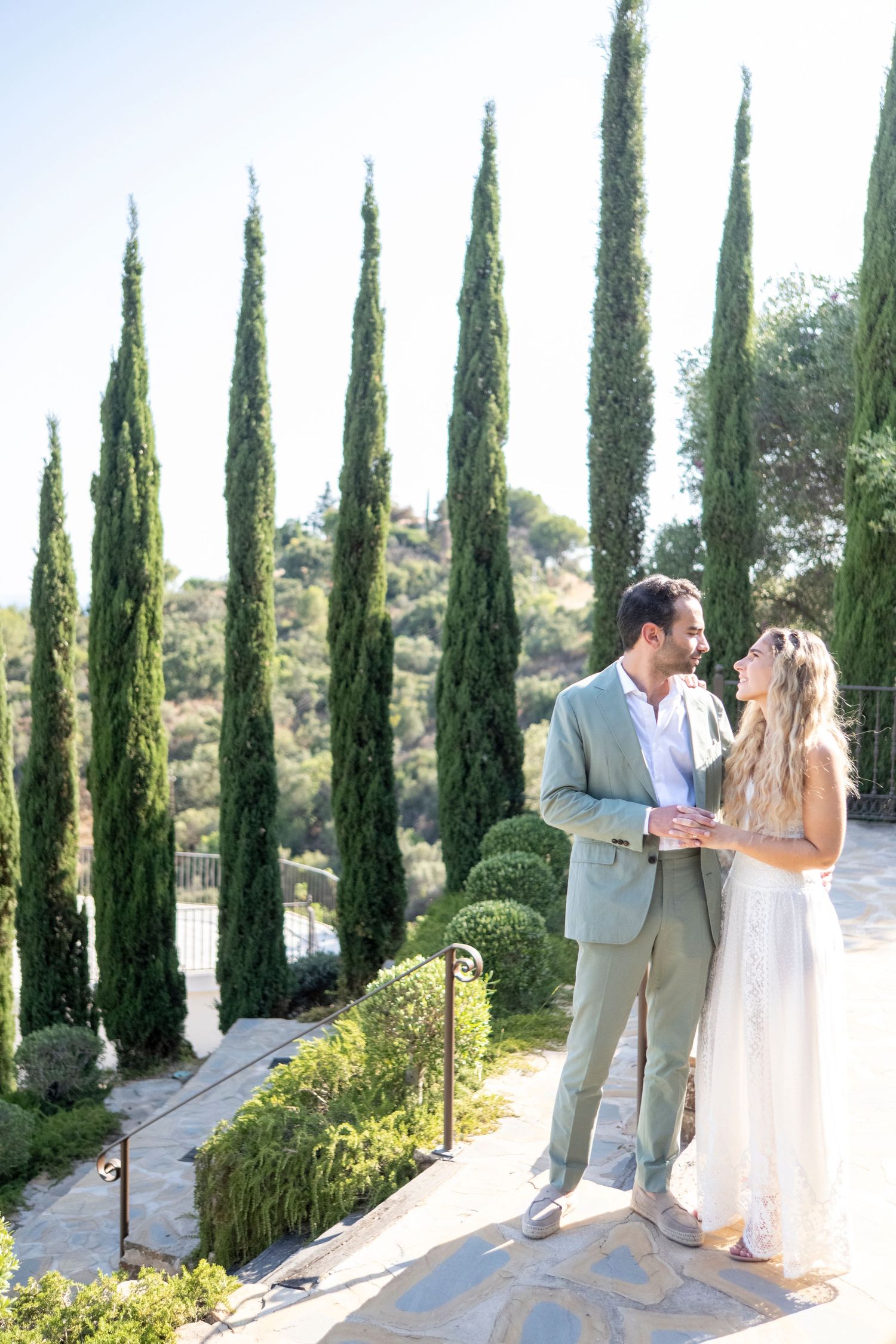 Romantic wedding couple embraces among tall cypress trees in a Mediterranean garden setting at sunset.