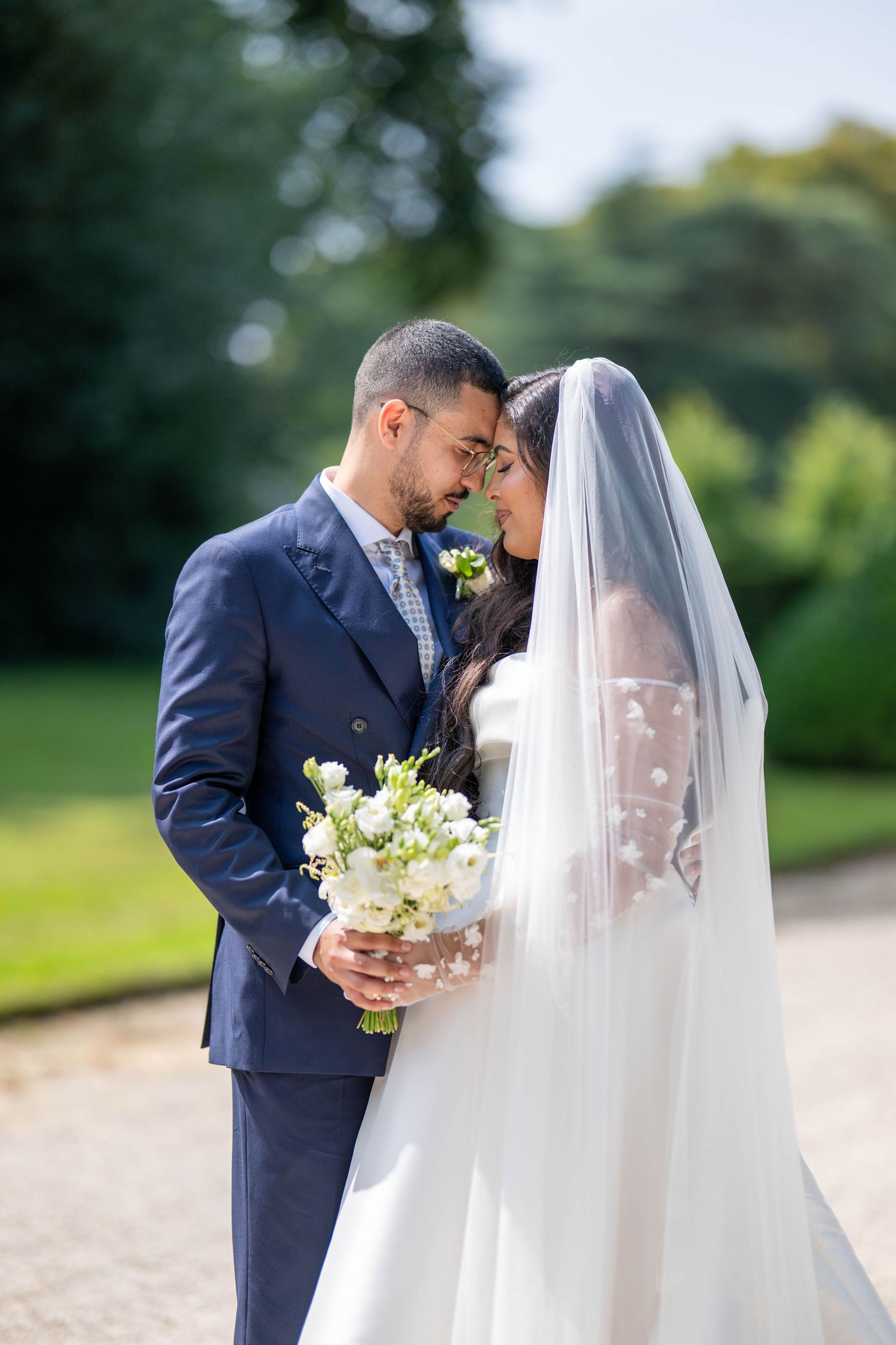 A romantic wedding couple shares intimate moments in a garden setting wearing navy suit and white veil.
