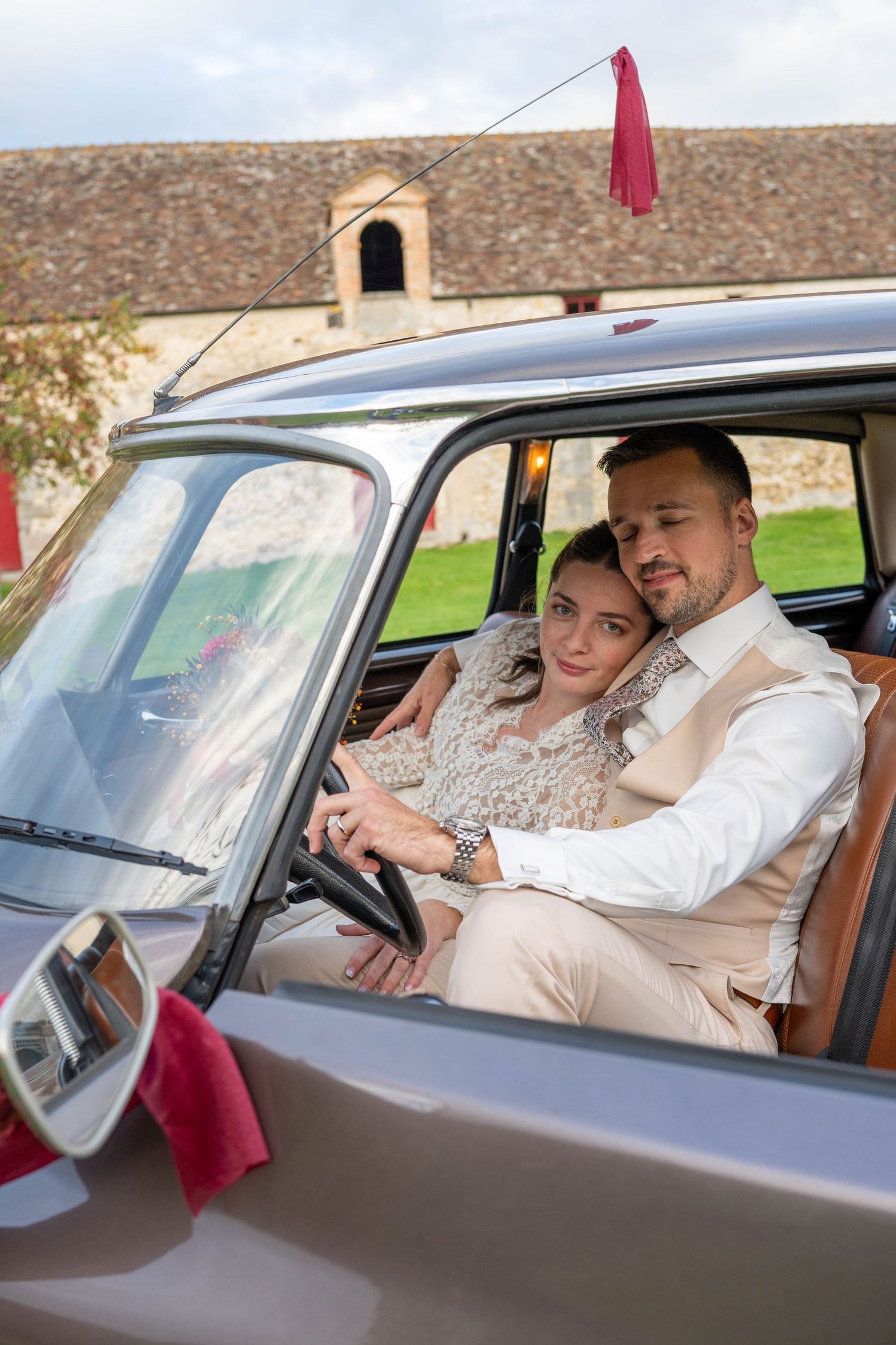 A couple sits in a vintage car decorated with red flags during their wedding celebration.