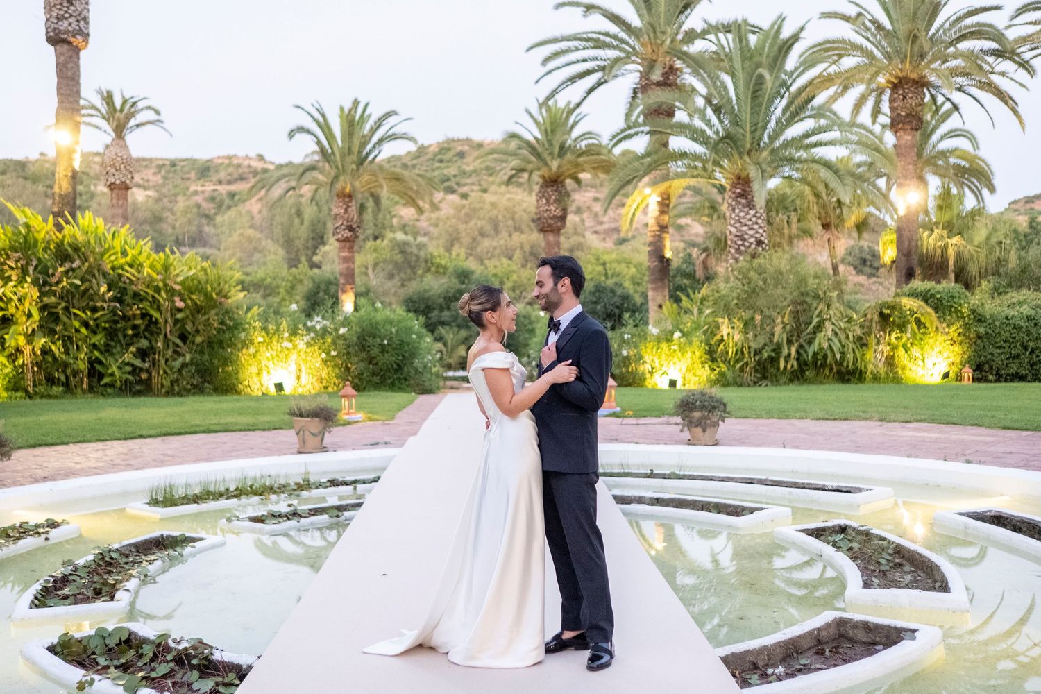 Couple in wedding attire embraces near a fountain in a lush garden setting at sunset.