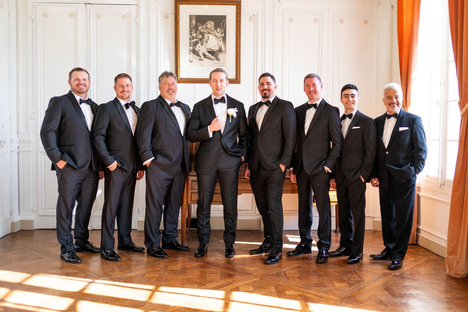 Groomsmen in black tuxedos stand in line against an elegant white wall at a formal wedding venue.