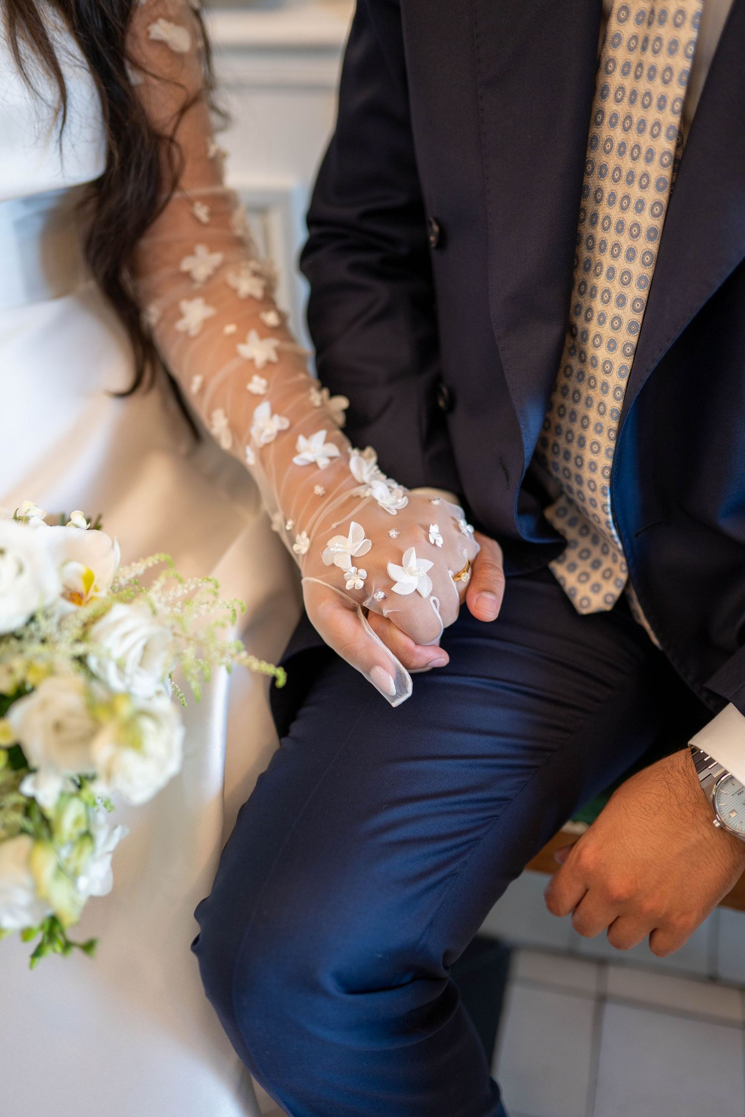 Close-up of wedding couple's hands holding each other, with bride wearing delicate lace sleeve and groom in navy suit.