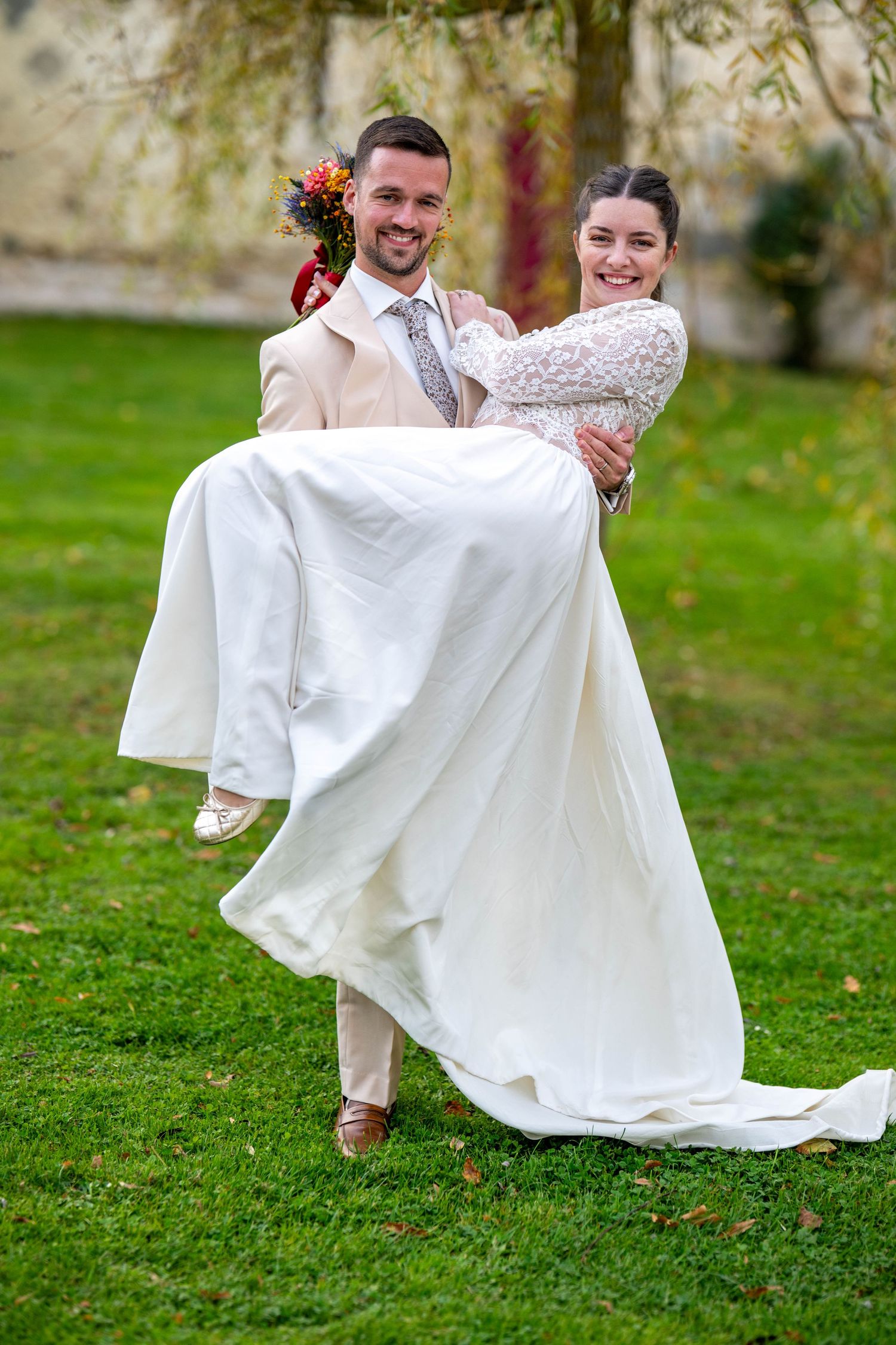 Groom lifting bride in flowing white wedding dress on grassy lawn.