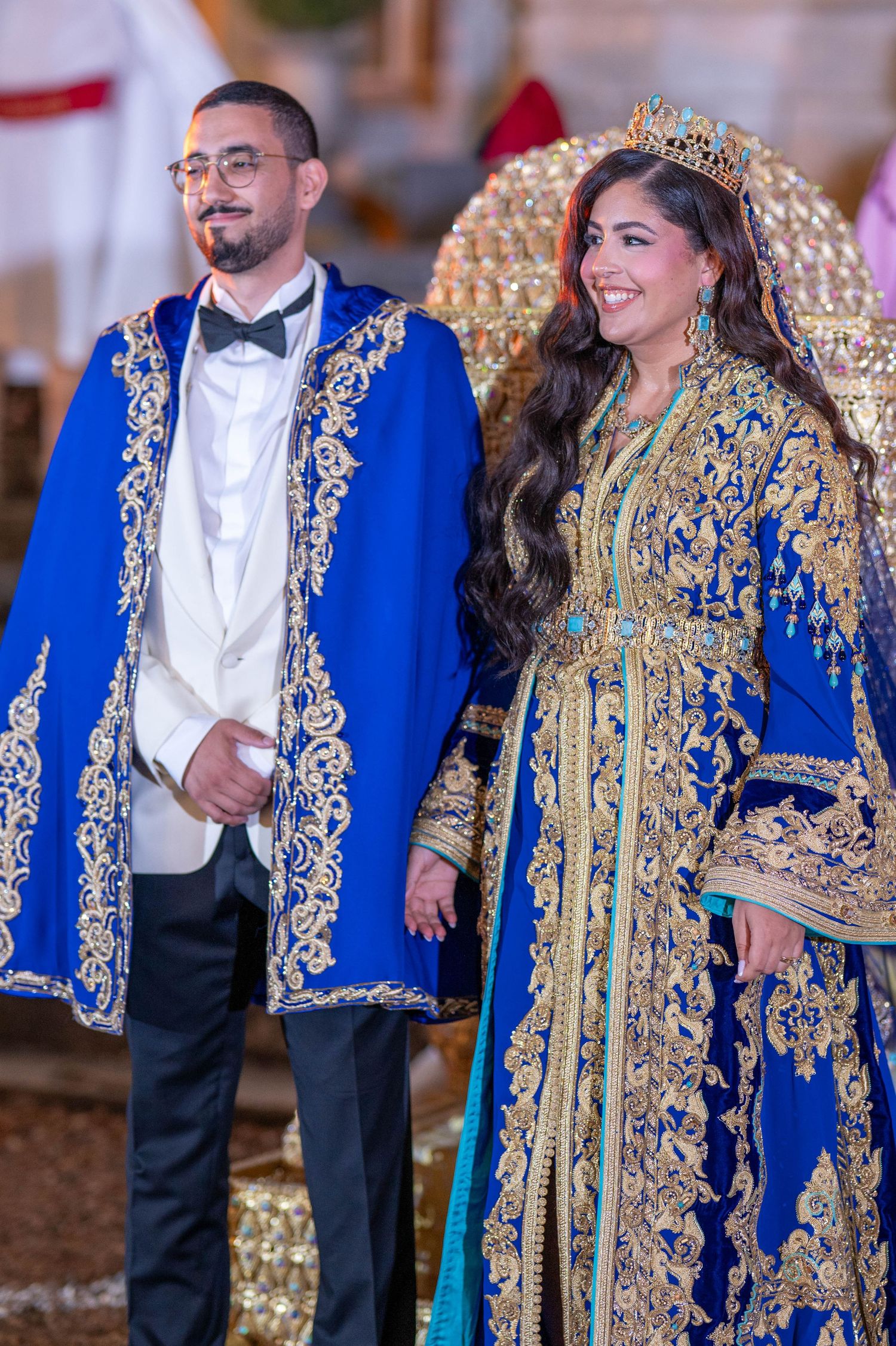 A couple wearing ornate royal blue and gold traditional formal attire pose together at a special celebration.