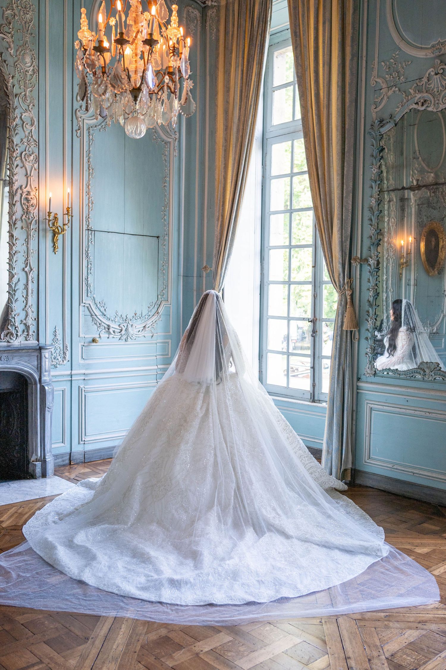 Elegant white wedding gown with long train displayed in ornate baroque room with crystal chandeliers and blue walls.