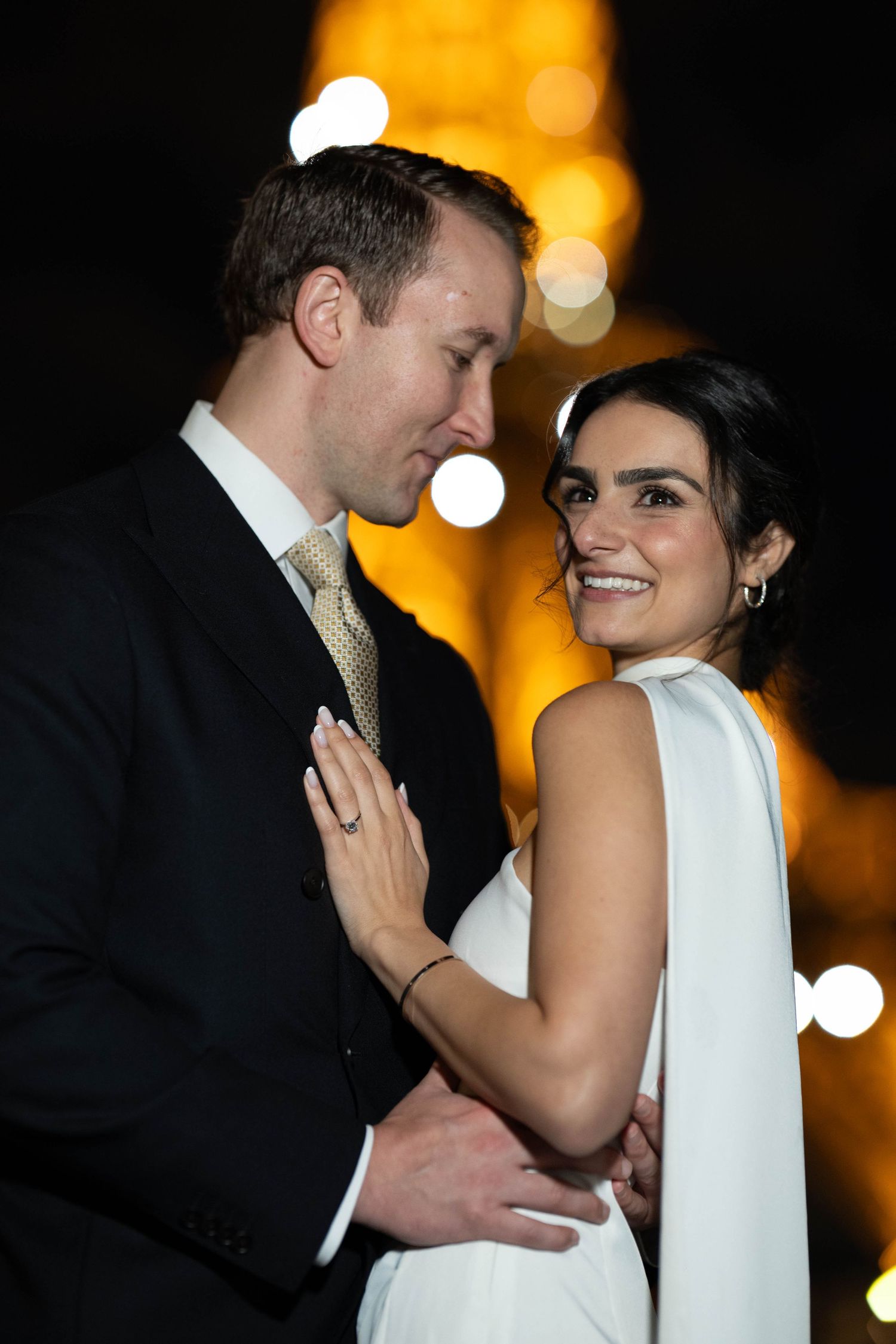 Romantic nighttime portrait of a couple in formal attire with glowing lights in the background.