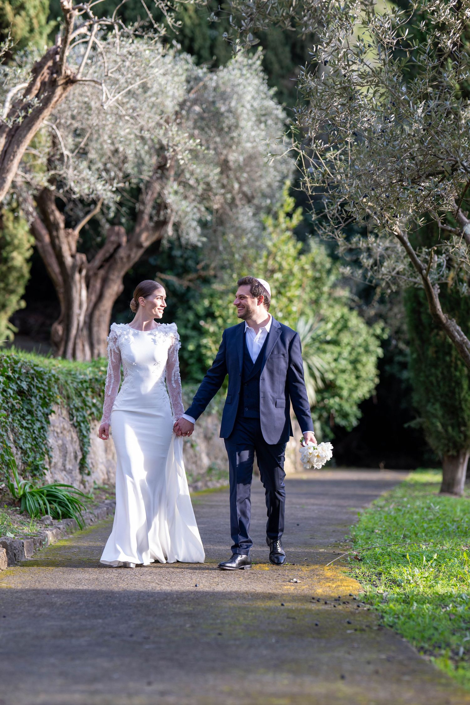 Newlyweds walking hand in hand along tree-lined garden path.