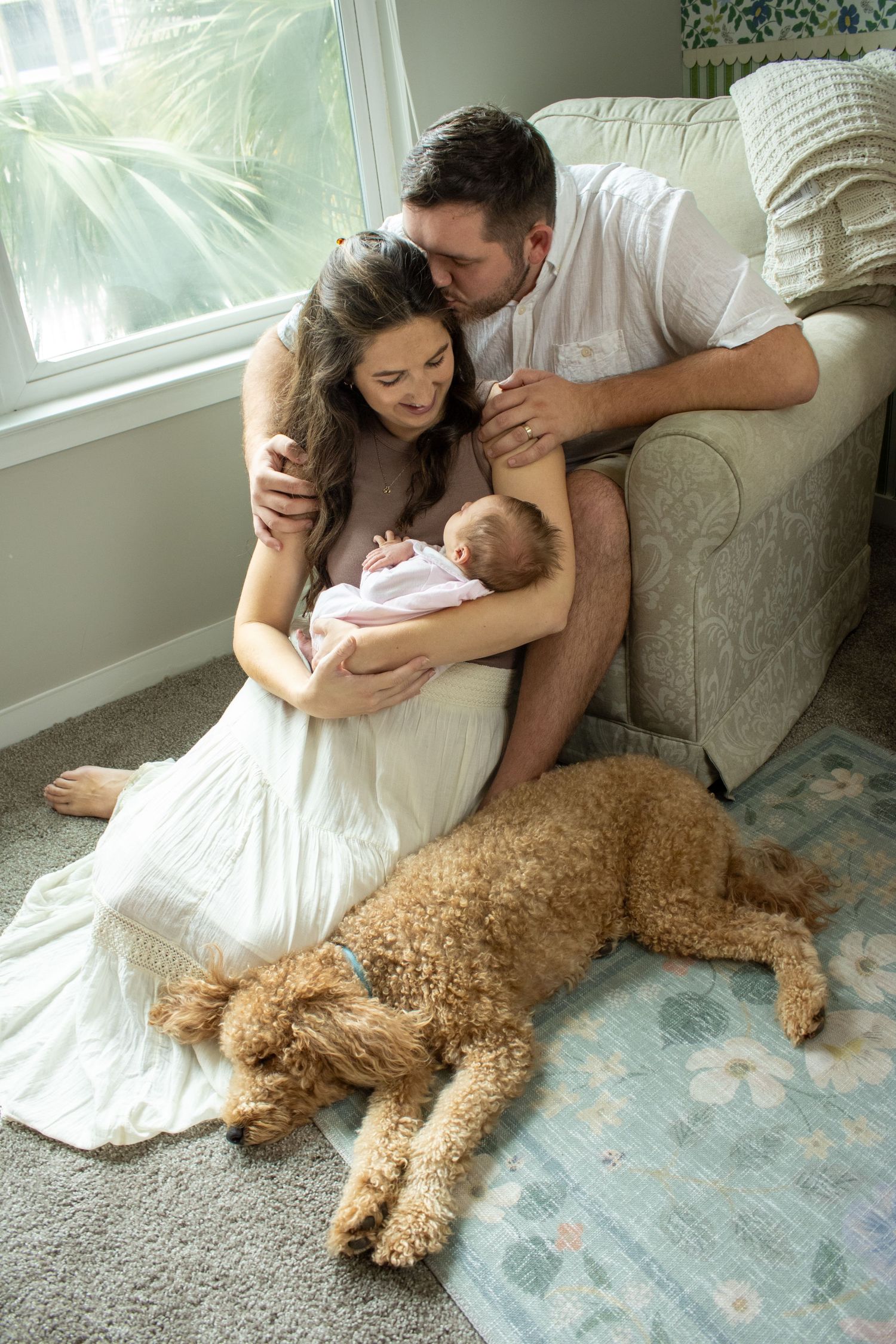 A couple sits on the floor beside their goldendoodle dog while cradling a newborn baby near a sunny window in Charleston South Carolina.