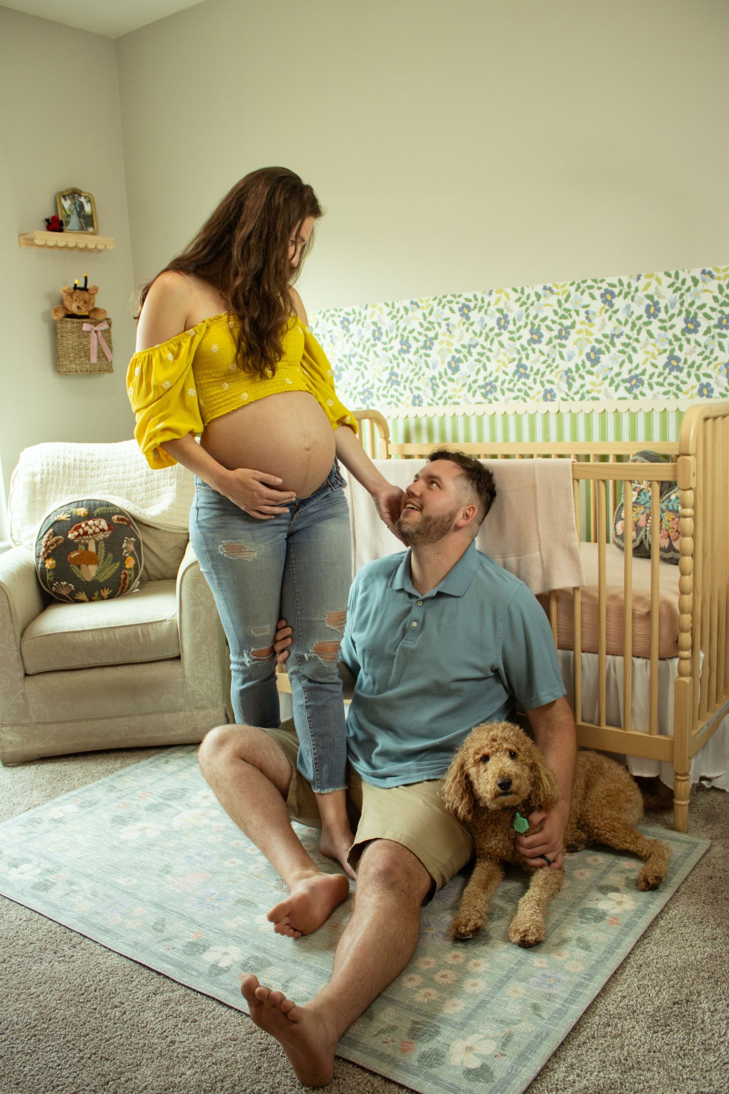 Couple sitting in nursery with their family dog as they prepare for new baby, with crib and yellow decor visible.