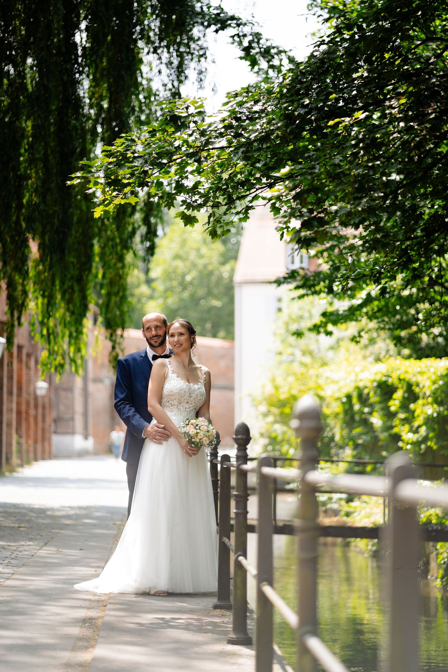 A couple shares a tender moment along a tree-lined pathway beside a peaceful waterway.