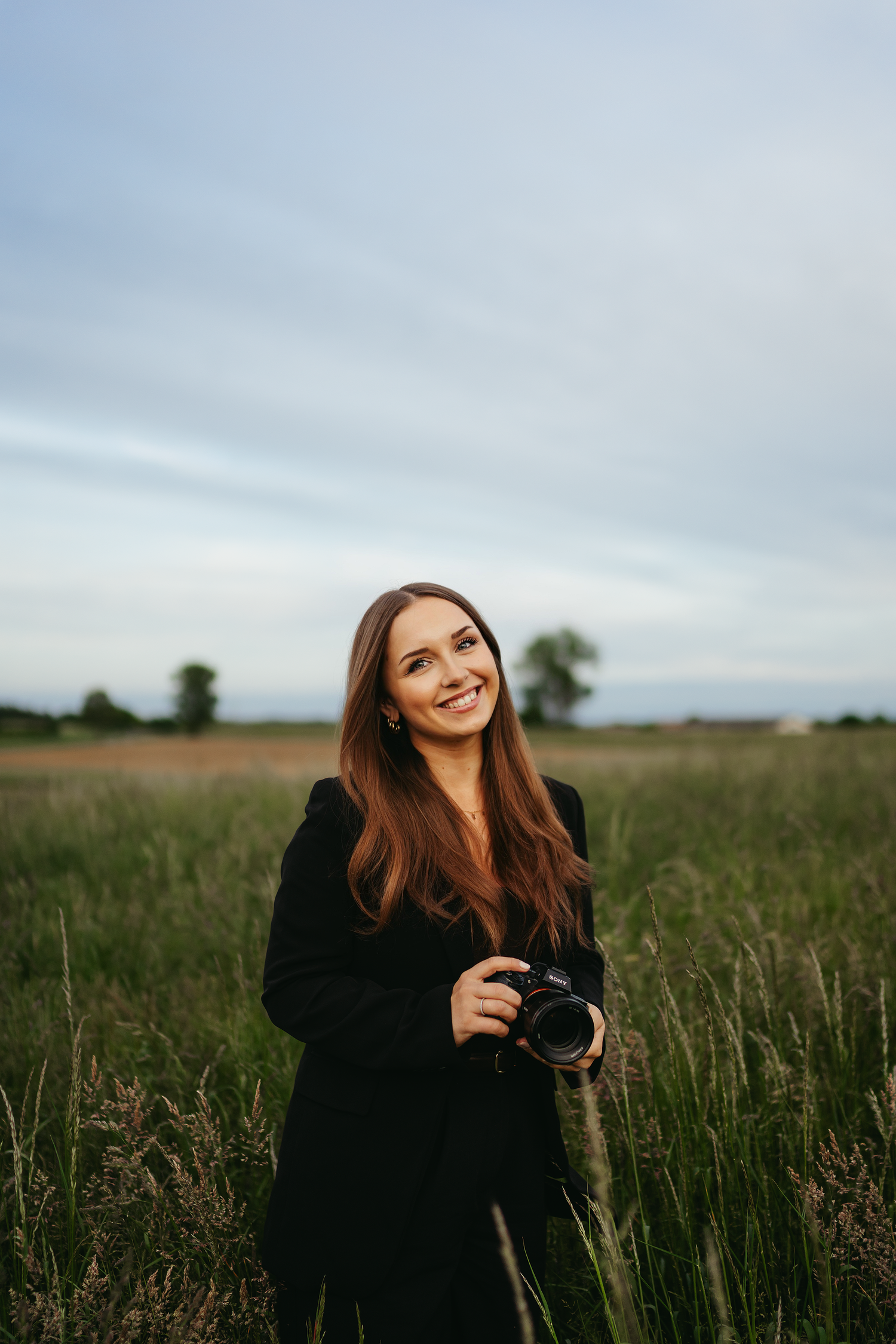 Photographer in black coat holding camera in field during golden hour.