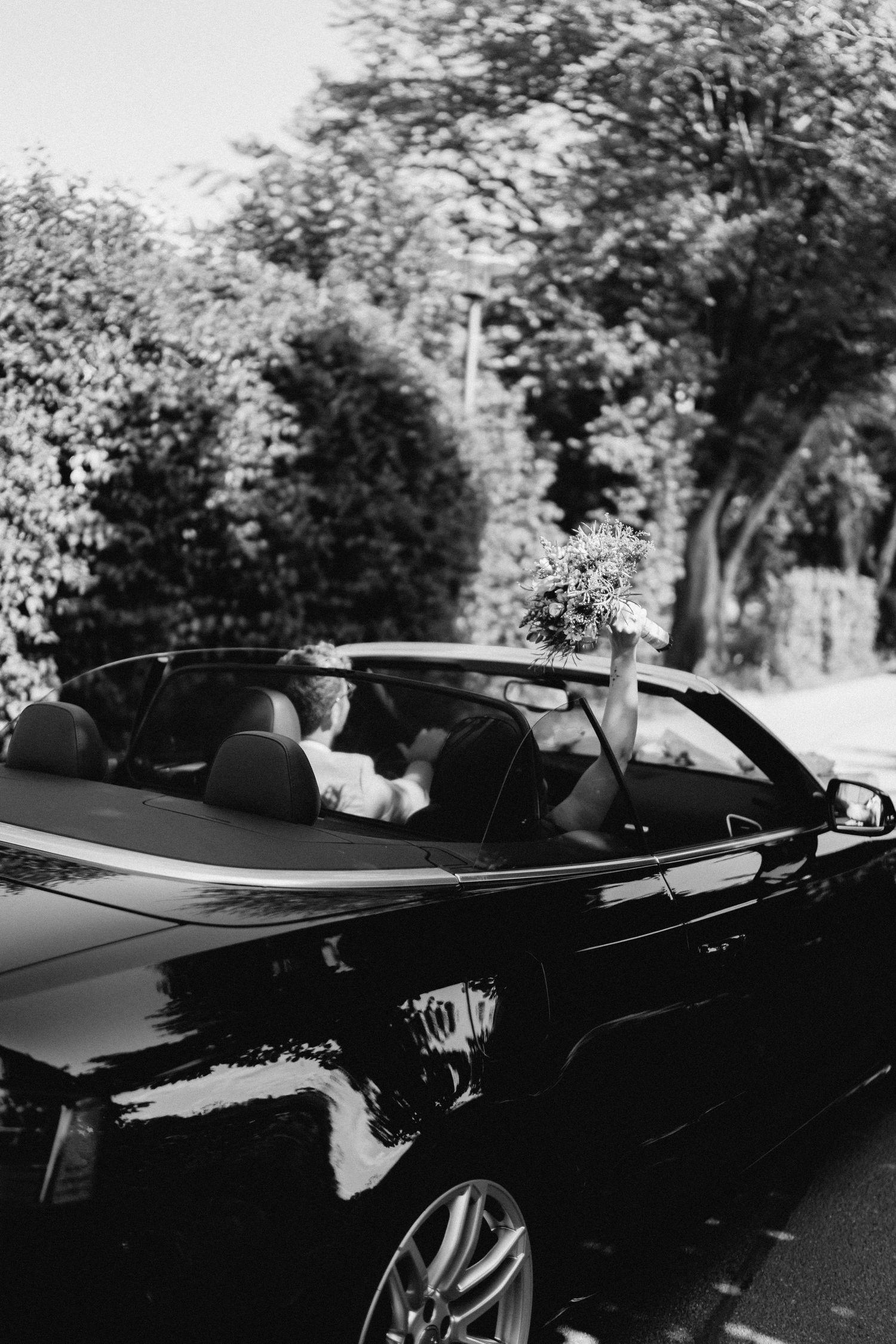 A sleek black convertible car parked under trees creates an elegant wedding day transportation scene.