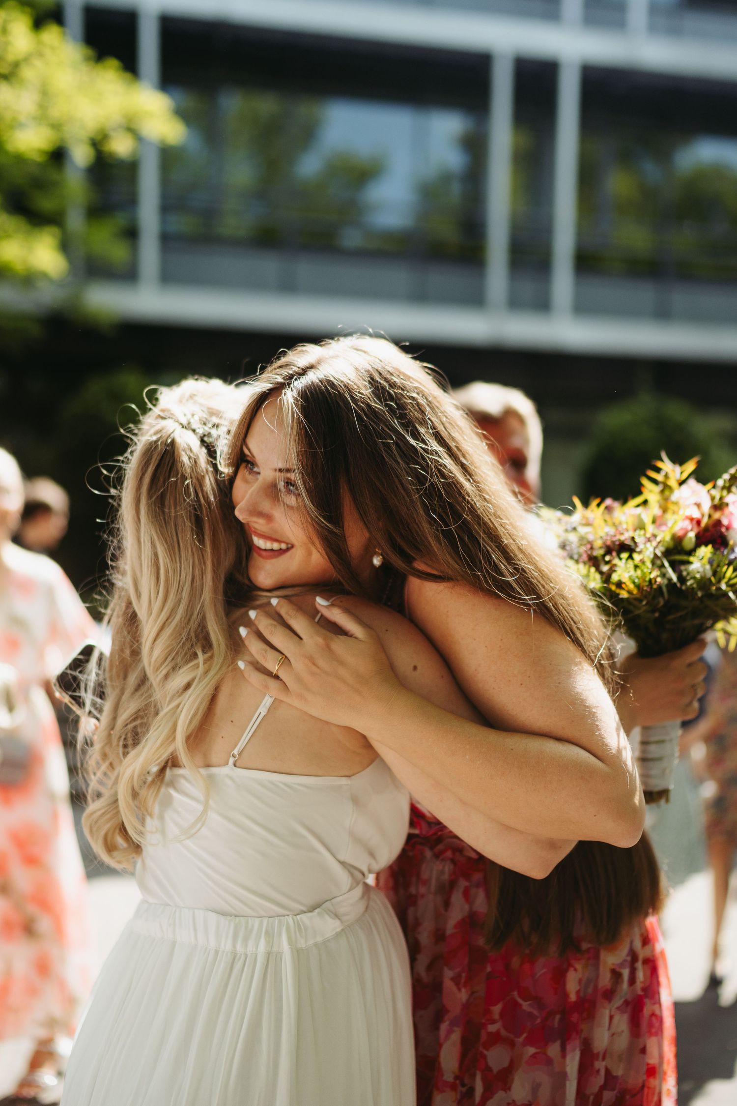 Wedding party members embrace in flowing white and burgundy dresses under sunlit trees.