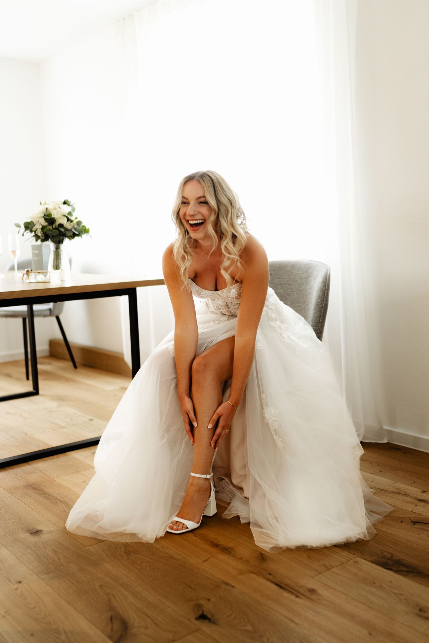 Bride in white gown adjusting shoes on hardwood floor in bright room.