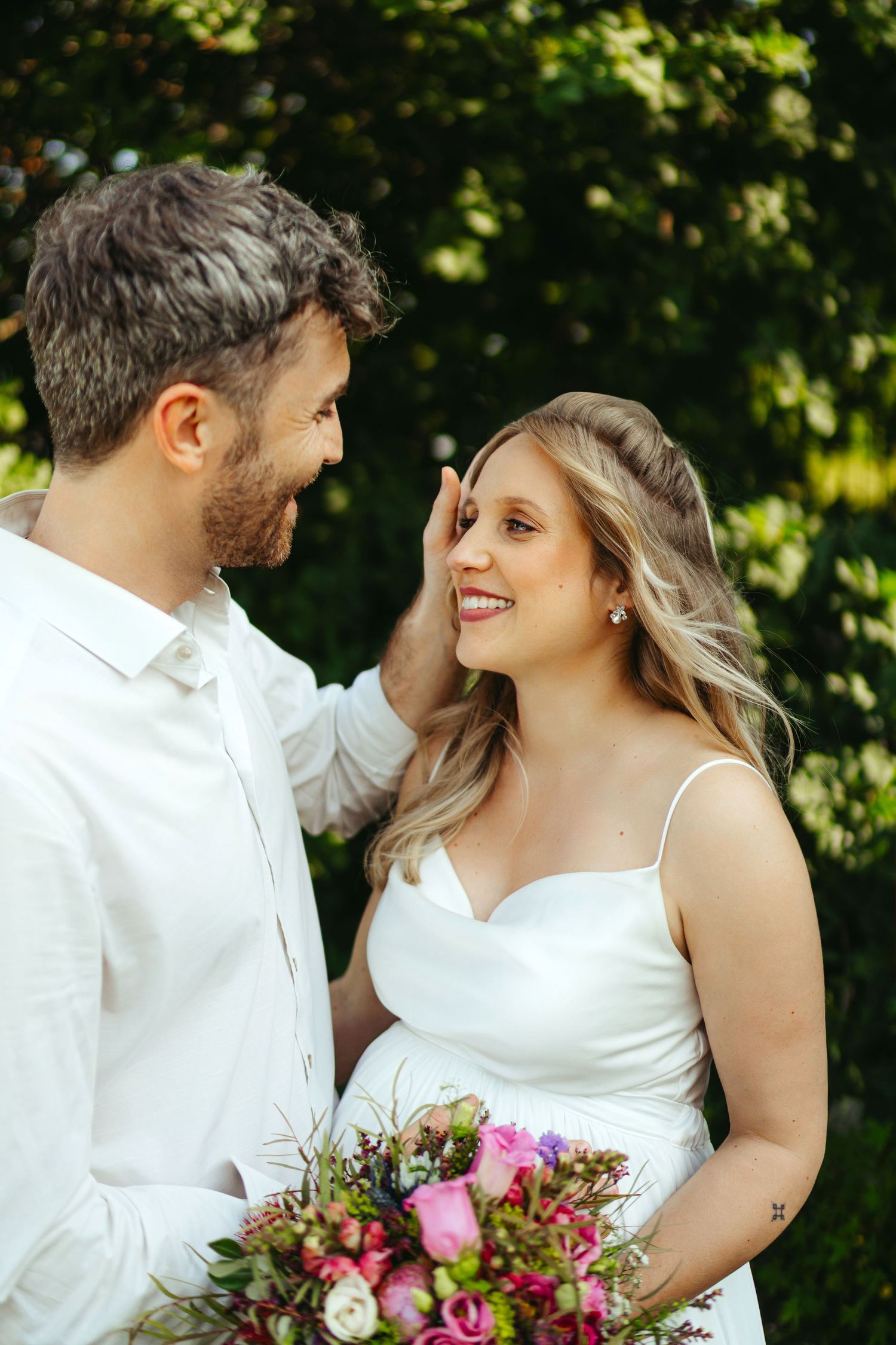 A couple in matching white wedding attire share intimate moments while holding a vibrant pink bouquet.