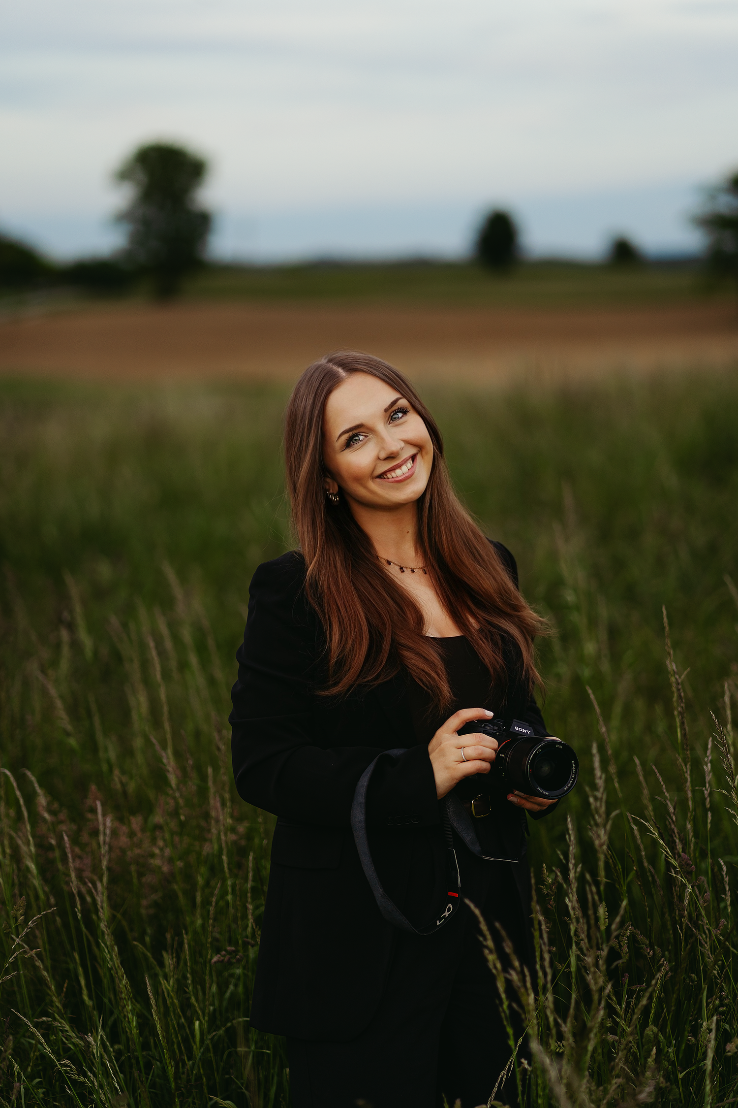 Professional photographer poses in a rustic field during golden hour with camera equipment.