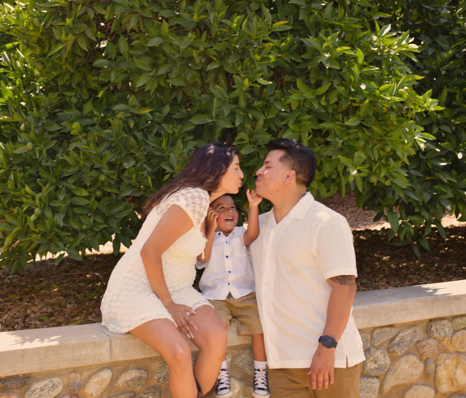 Family dressed in white sitting on stone wall under green foliage.