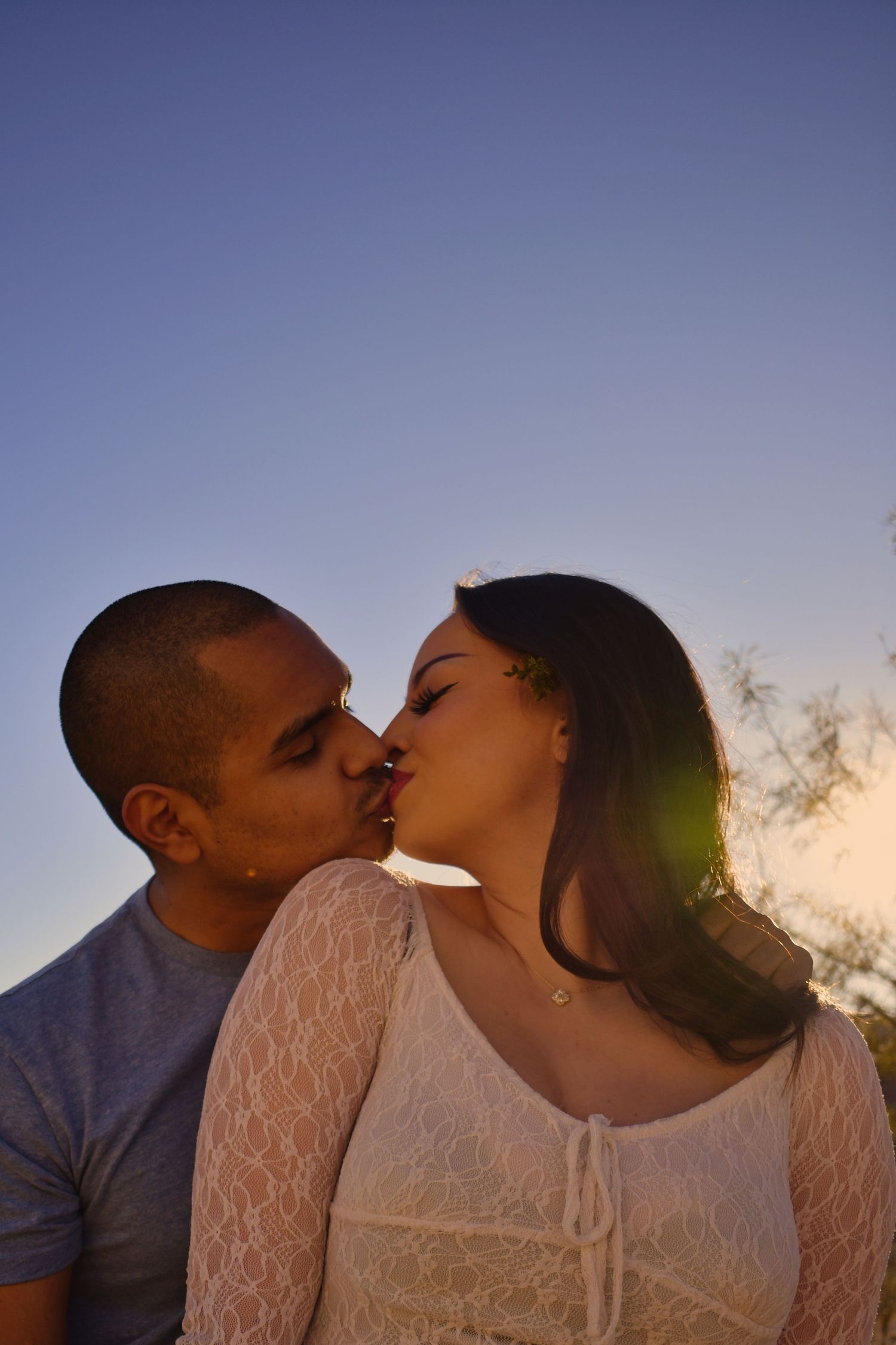 A romantic couple sharing a tender kiss at sunset with sunlight creating a warm backlit glow against a blue sky.