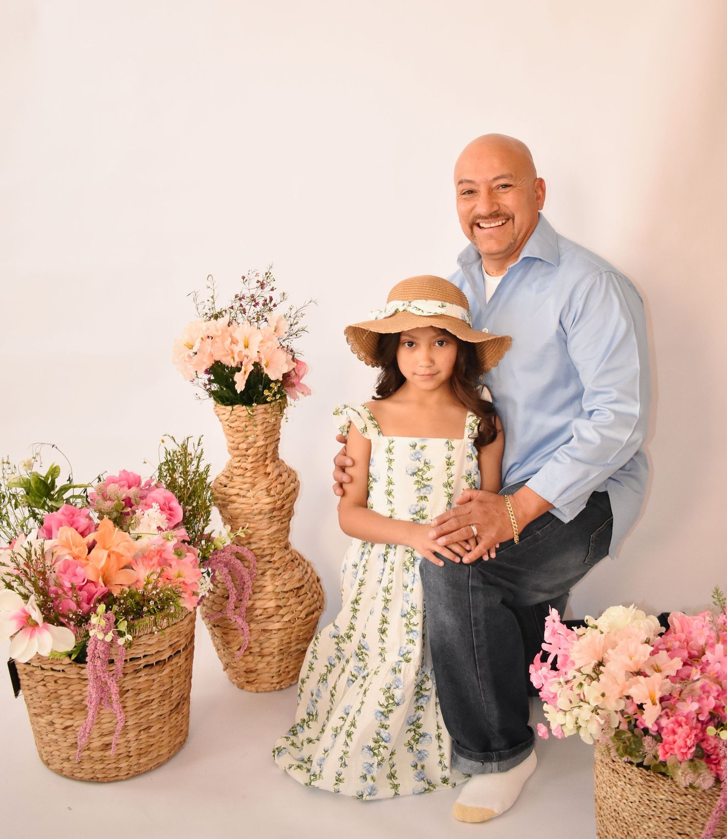 Man and young girl in floral dress and hat pose together among pink flower arrangements in wicker baskets.