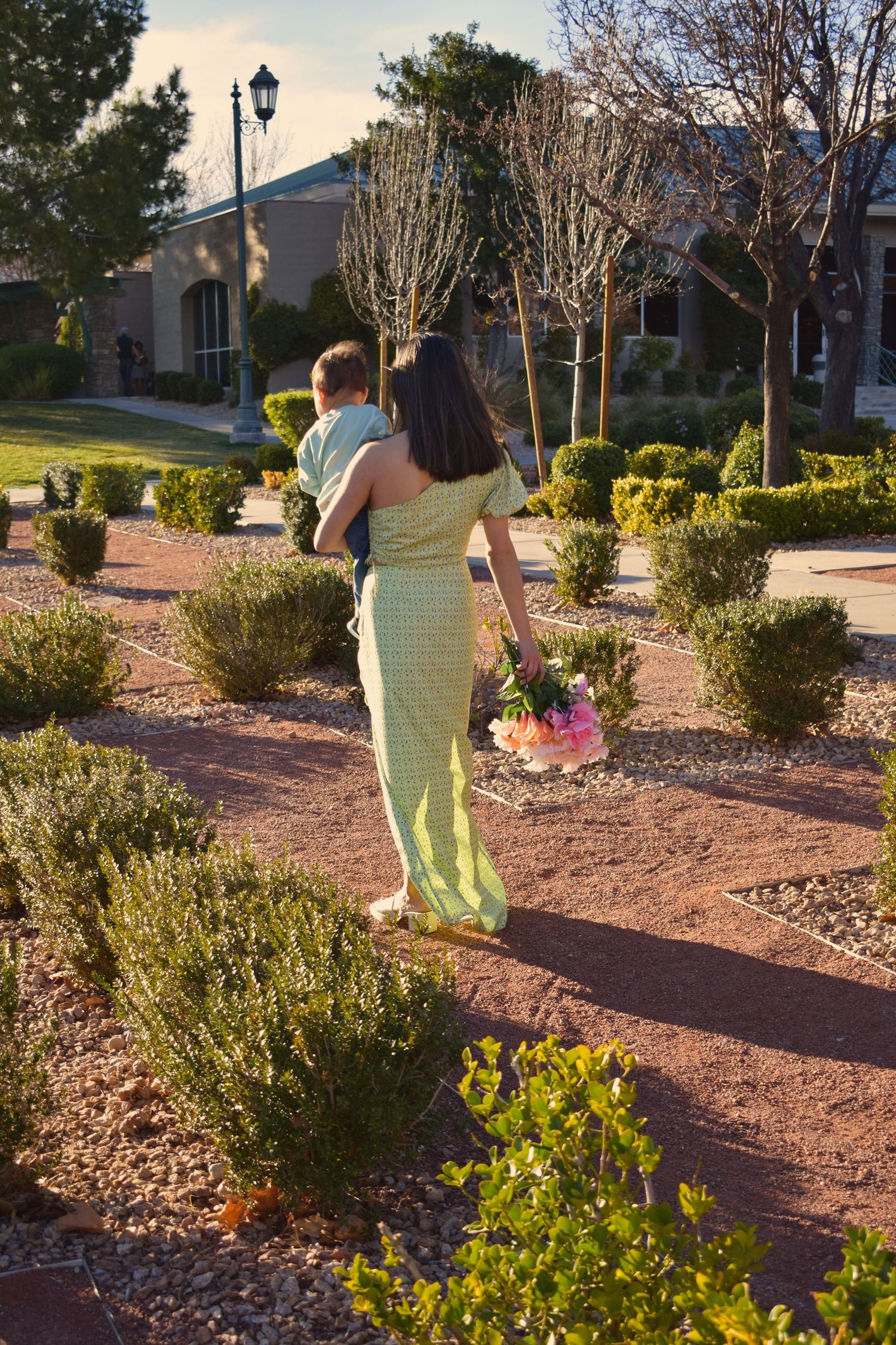 Two people walk along a landscaped garden path with ornamental shrubs and a lamp post at sunset.