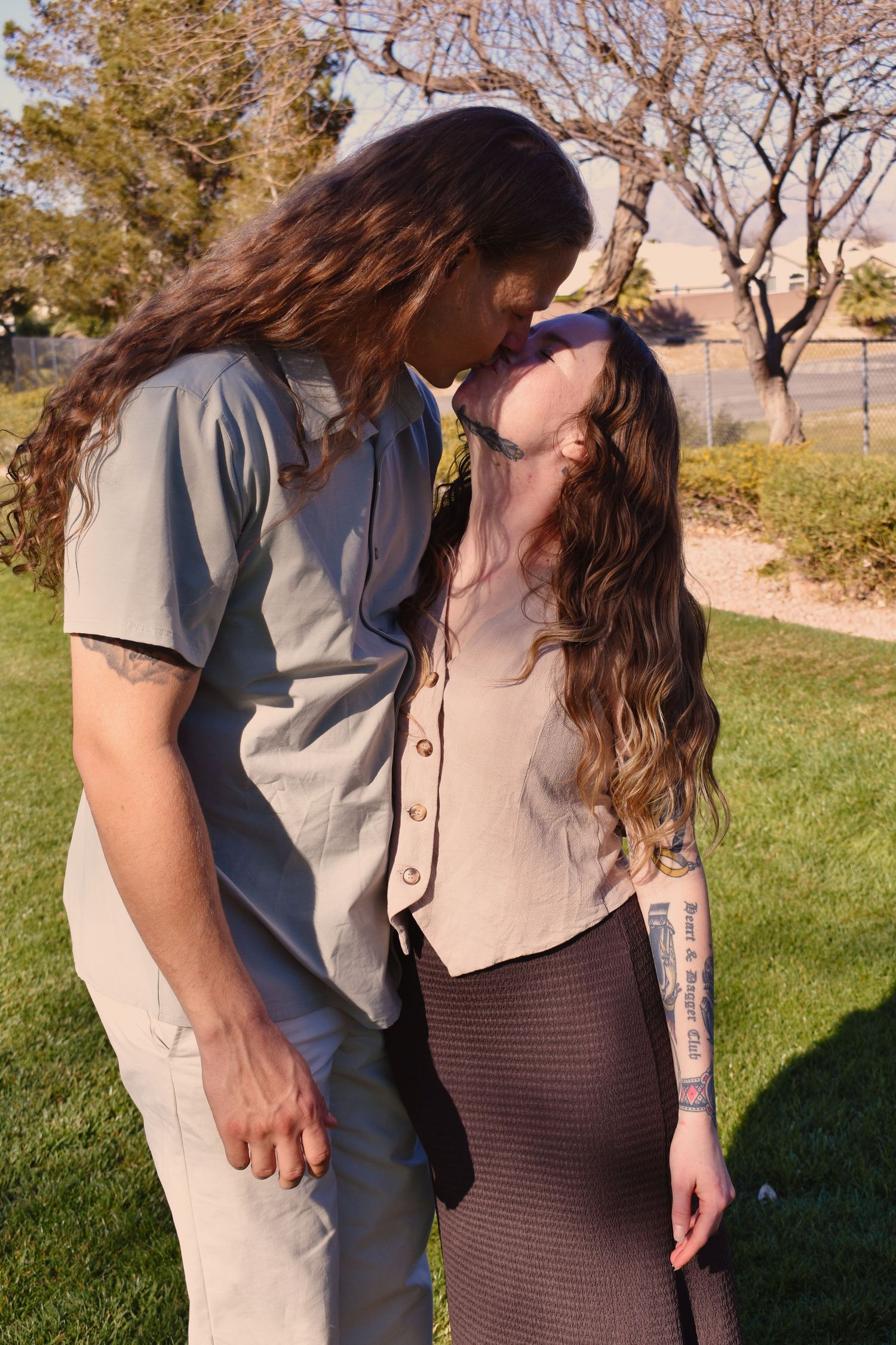 A couple embraces tenderly in a garden setting with sunlight casting shadows on the grass.