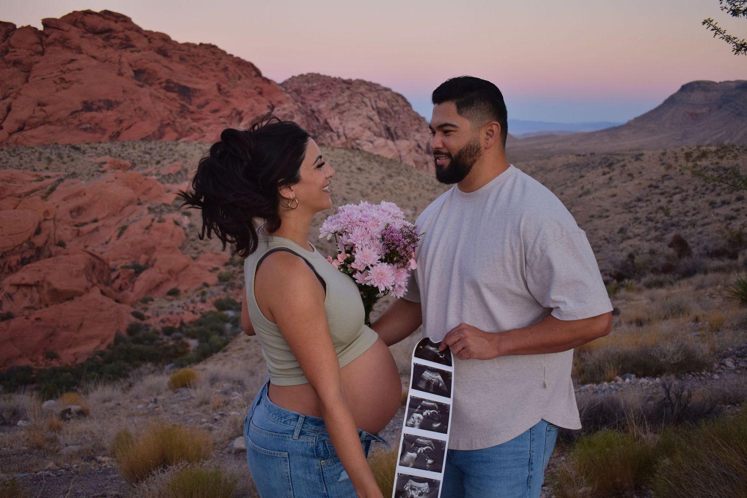 A couple shares a tender moment holding pink flowers against desert red rocks at dusk.