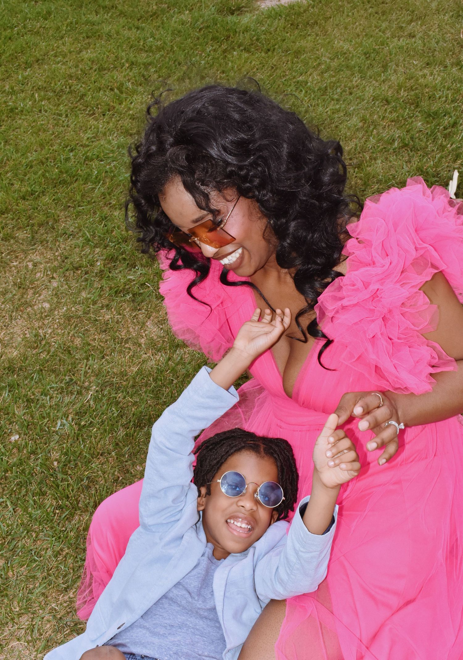 A woman in a pink ruffled dress laughs with a child wearing blue sunglasses on a grassy lawn.