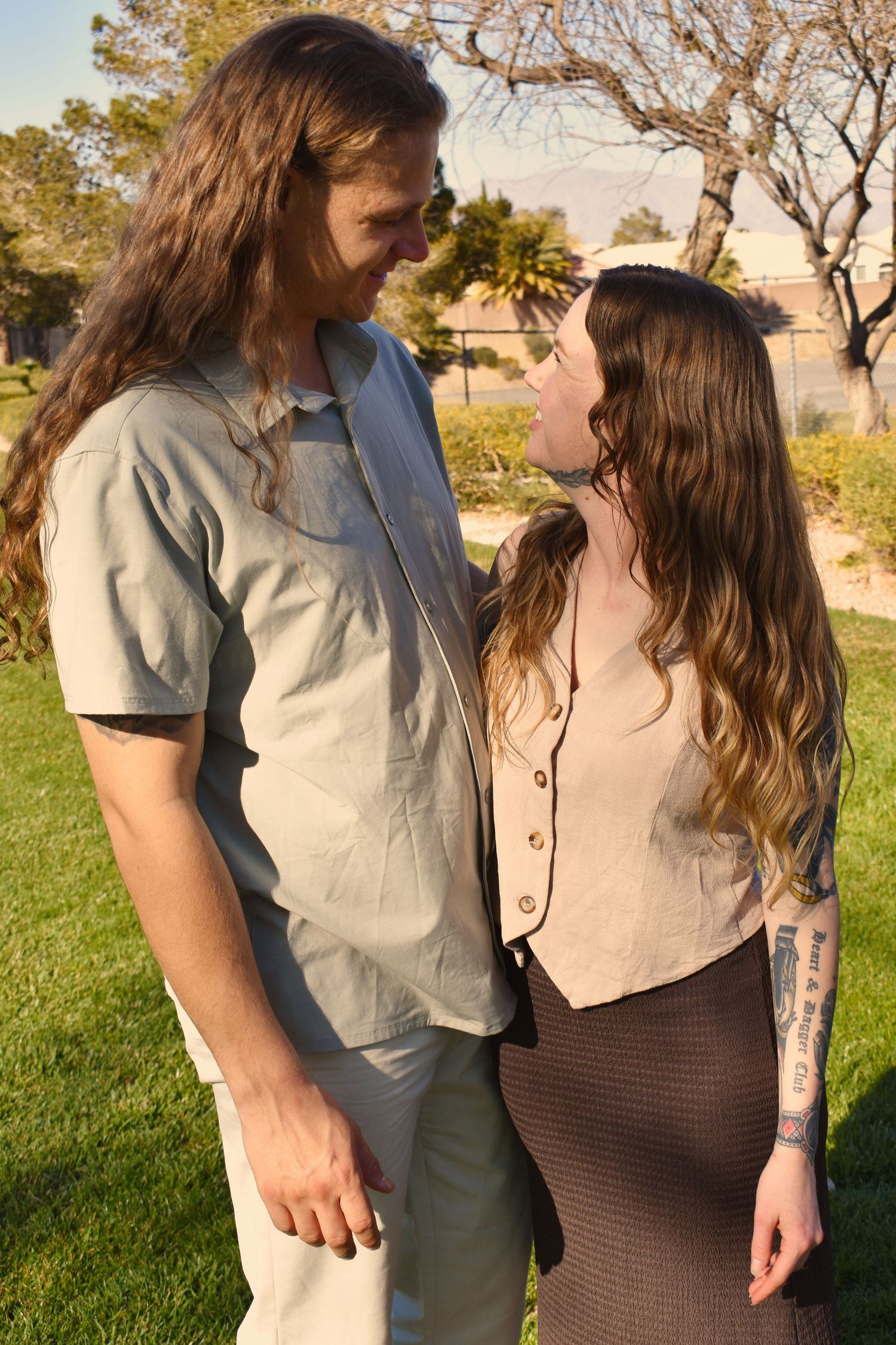 A couple in casual attire embraces outdoors against a natural backdrop during golden hour.