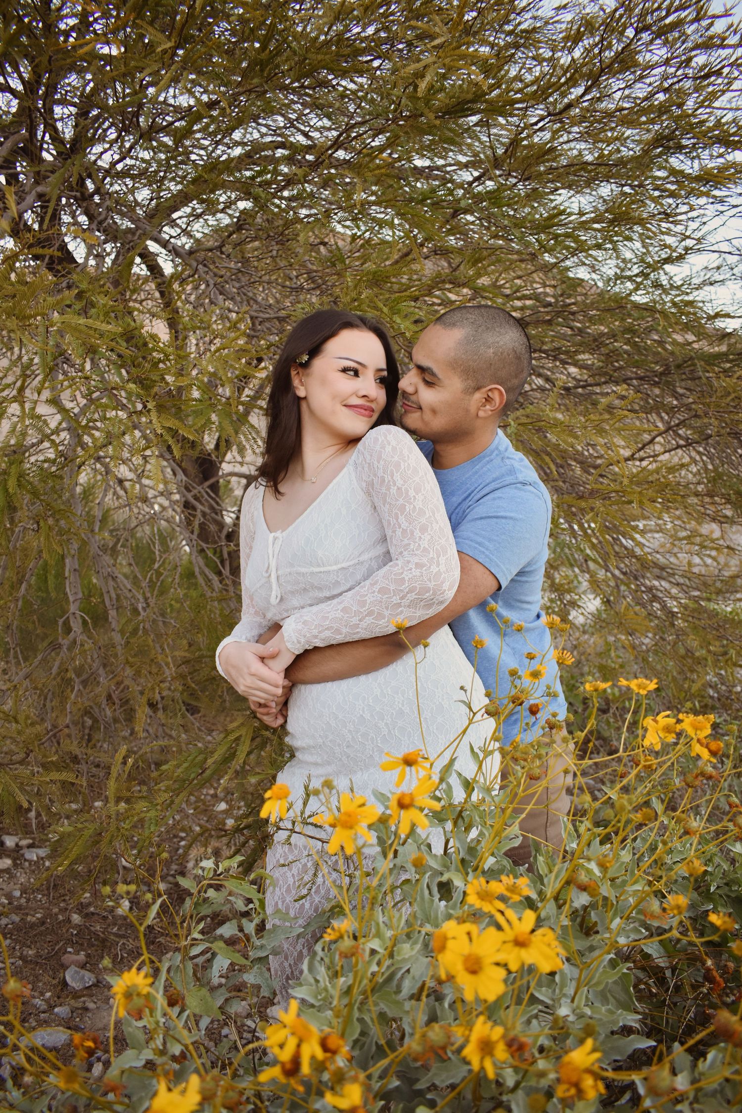 A romantic couple embraces among yellow wildflowers and desert brush during a sunset photoshoot.