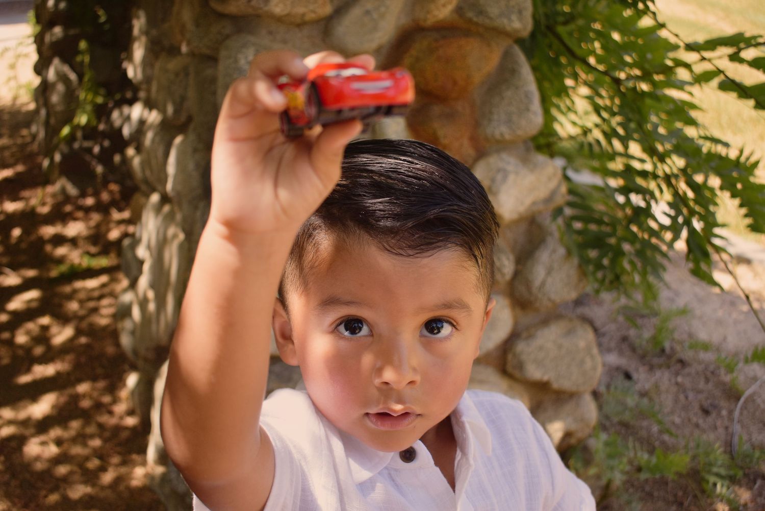 Child holding red object while sitting outdoors near stone wall.