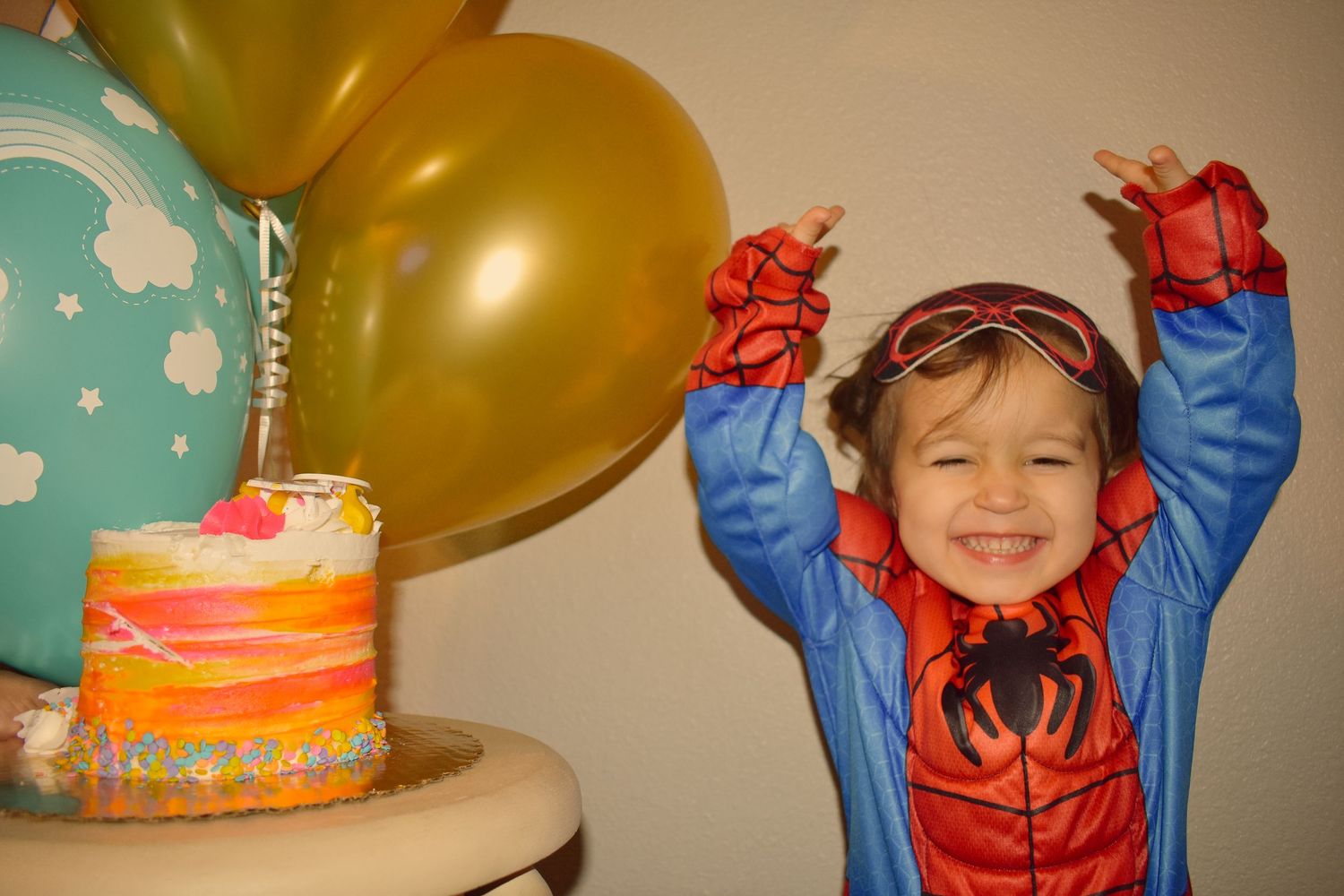 Child in Spider-Man costume celebrates with gold balloon and colorful birthday cake.