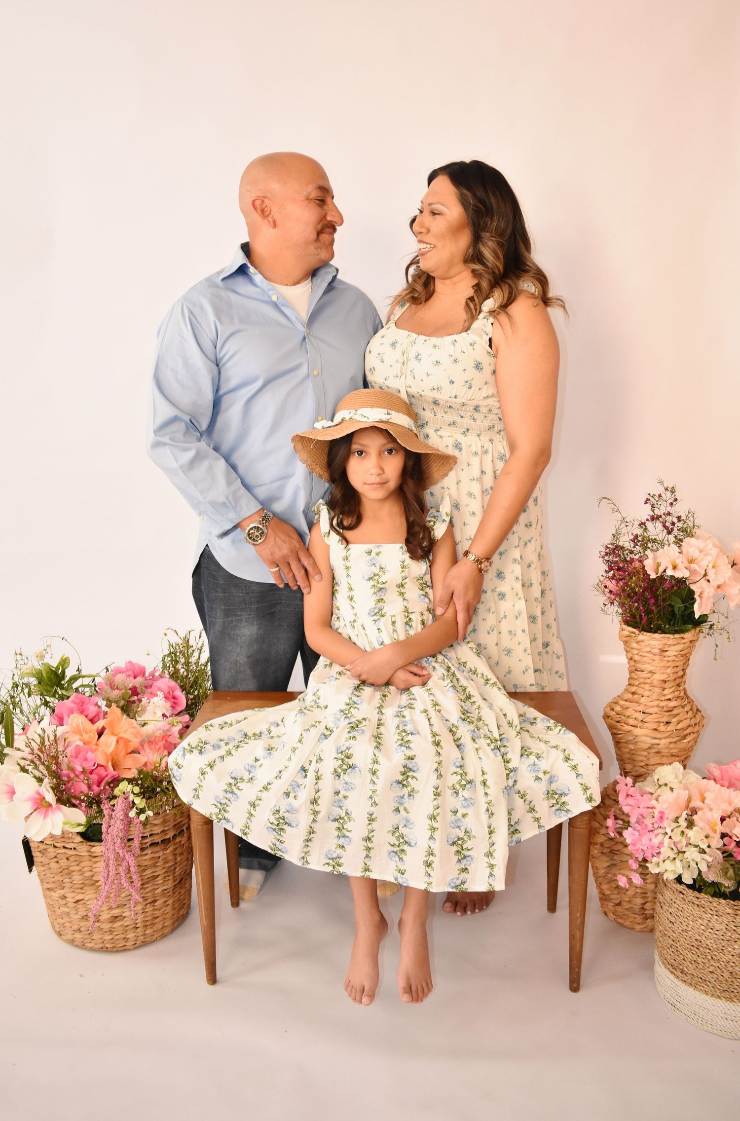 A family of three poses in a studio with floral arrangements, parents in matching outfits with daughter in a white floral dress.