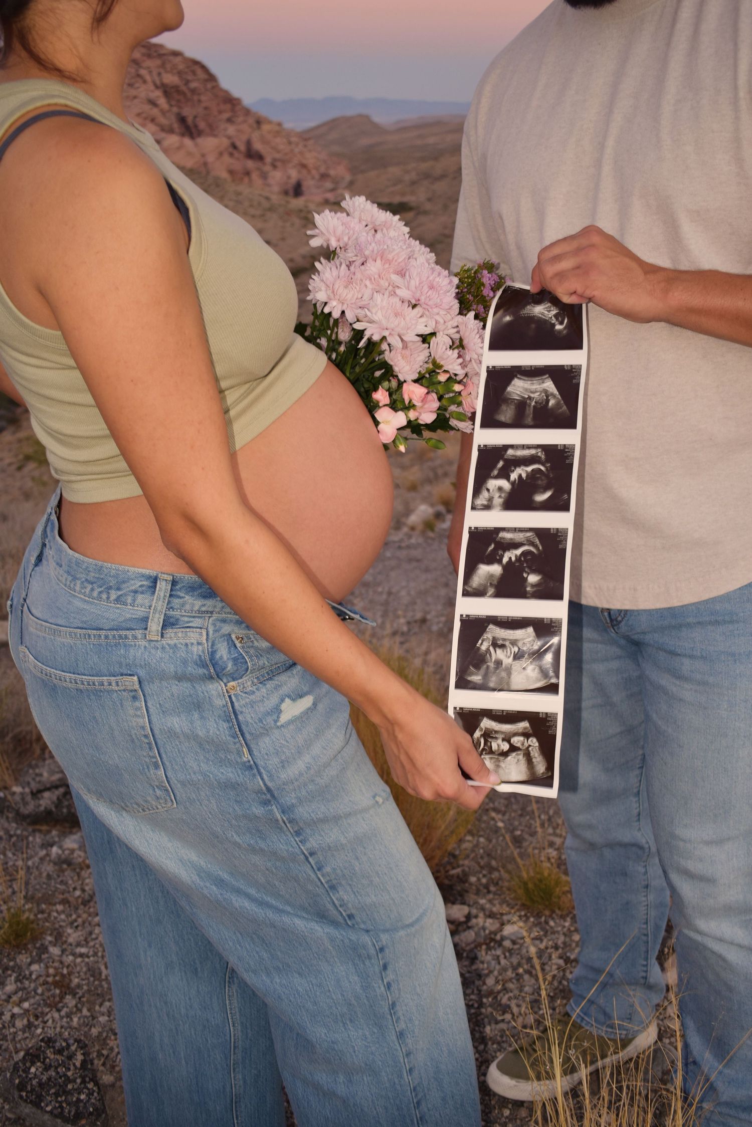 A couple holds ultrasound photos while standing in a desert setting with pink flowers.