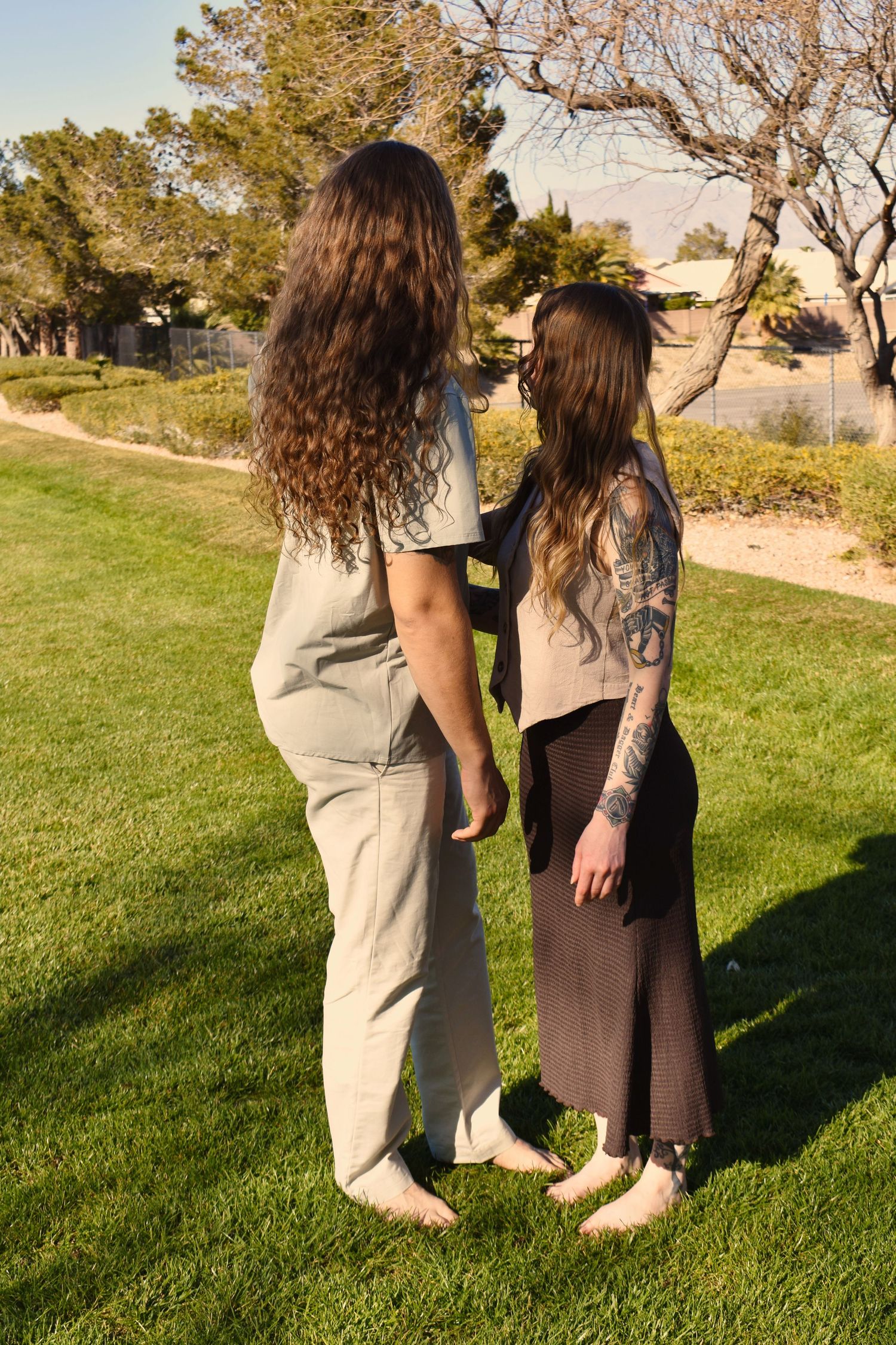 Two people in casual attire standing together on a grassy hill during sunset casting long shadows on the ground.