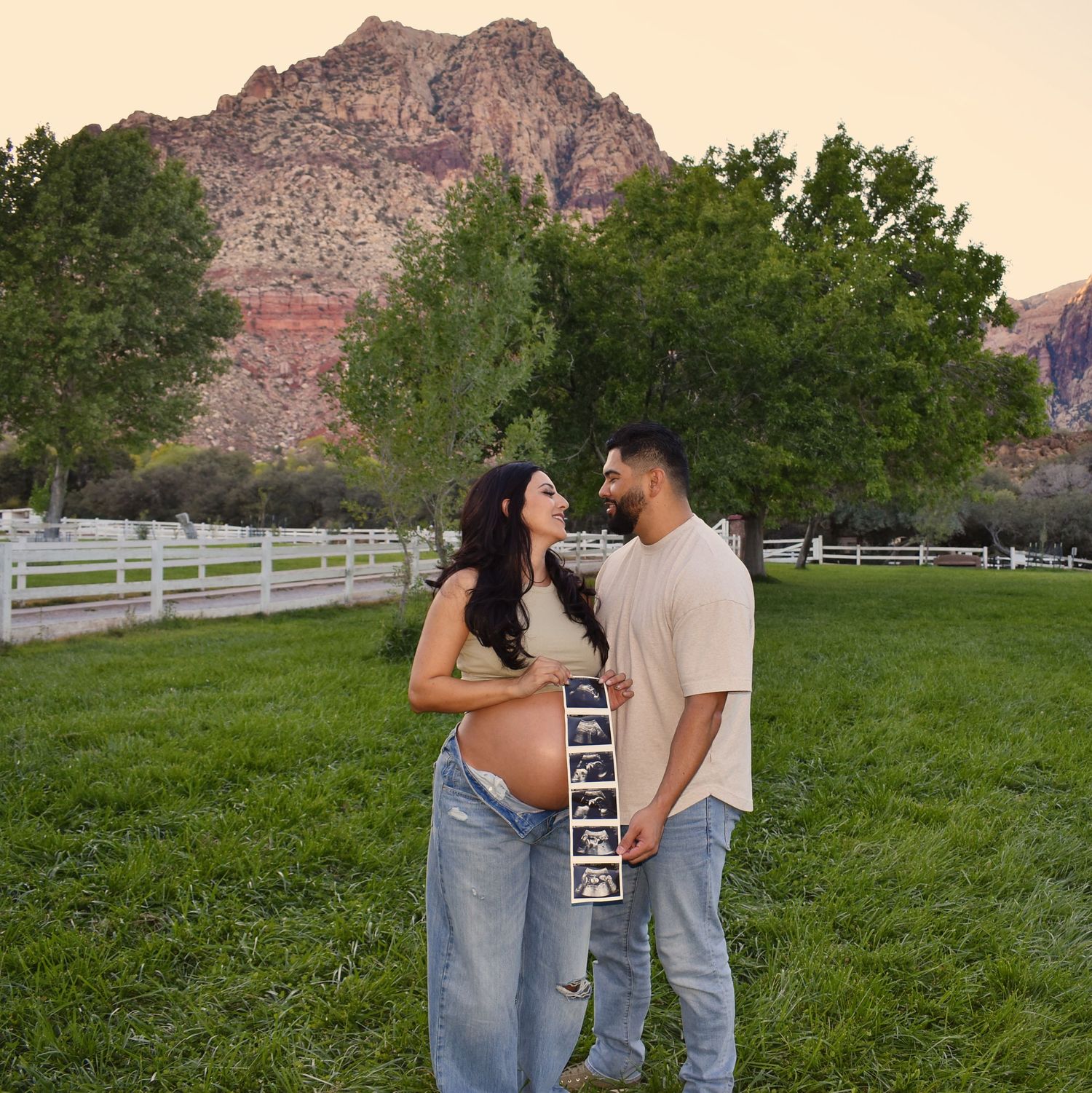An expectant couple poses with ultrasound photos in front of mountain scenery and white fencing.