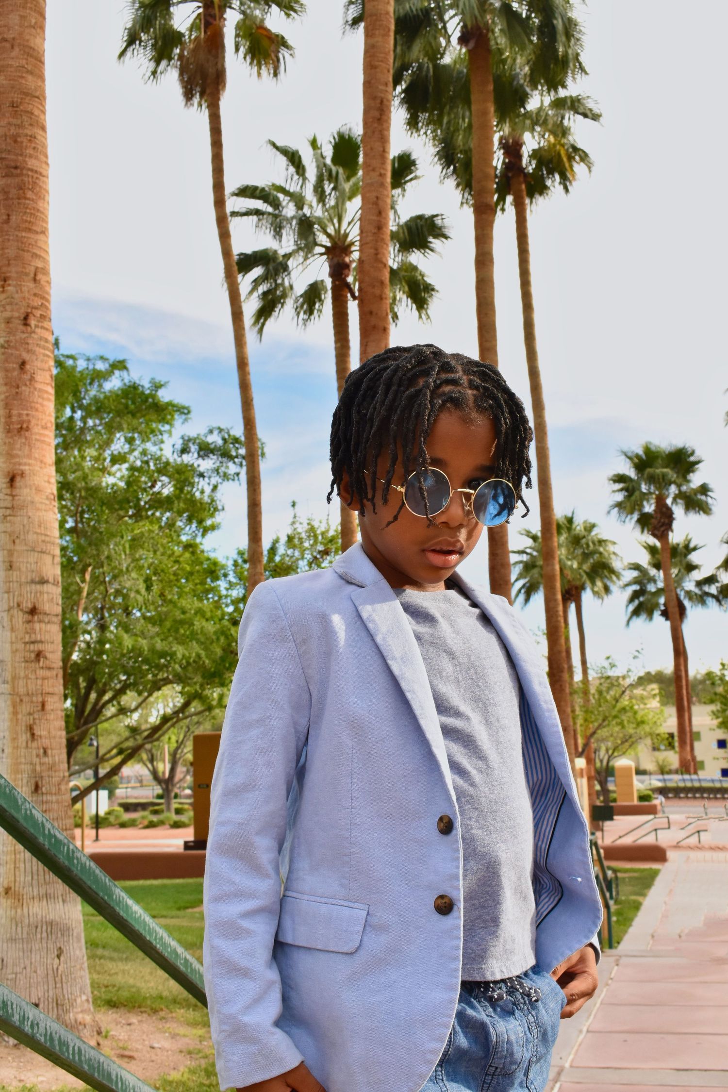 Young person in blue blazer and round sunglasses poses outdoors among tall palm trees on a sunny day.