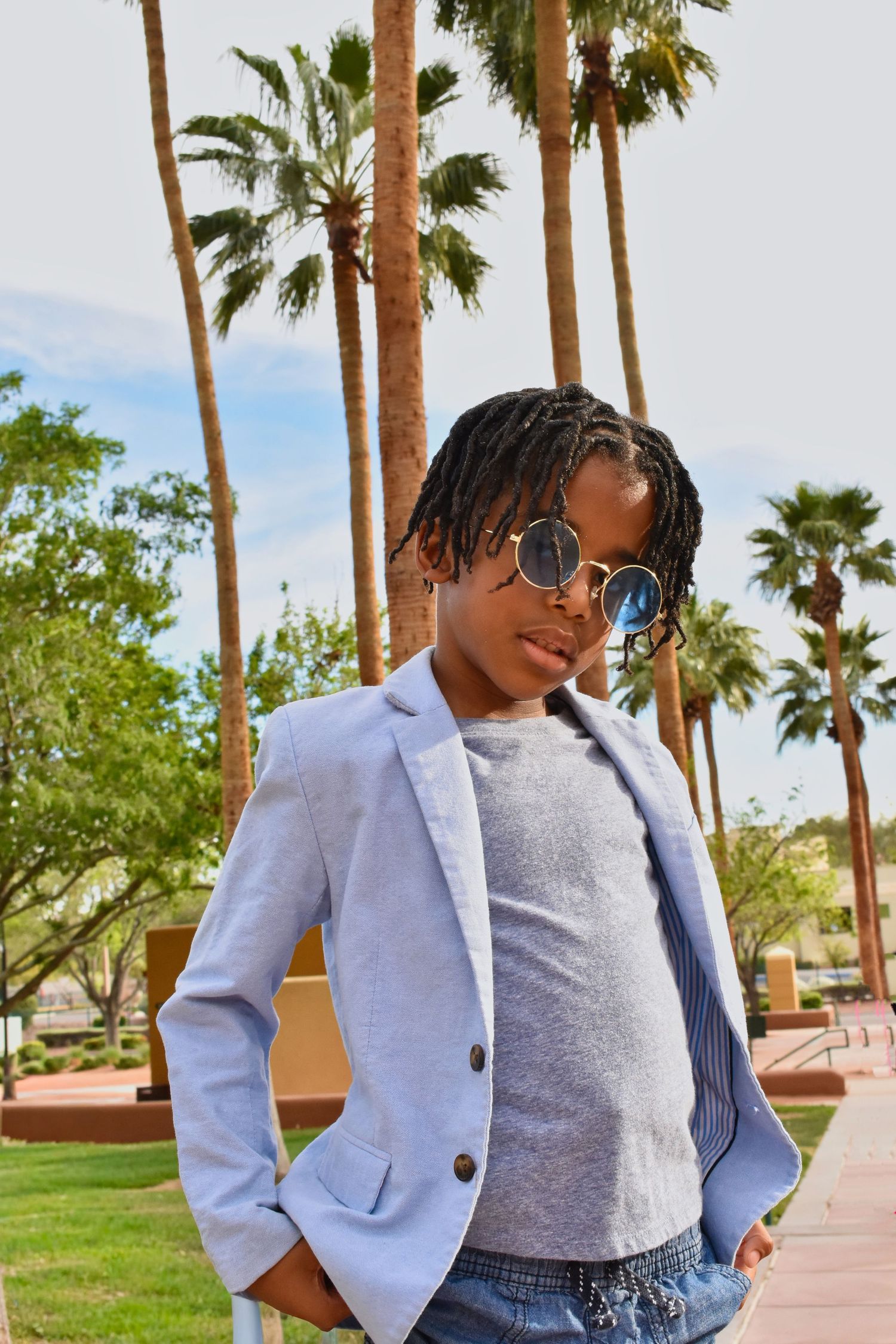 Stylish young person wearing round sunglasses and a light blue blazer poses outdoors with palm trees in the background.
