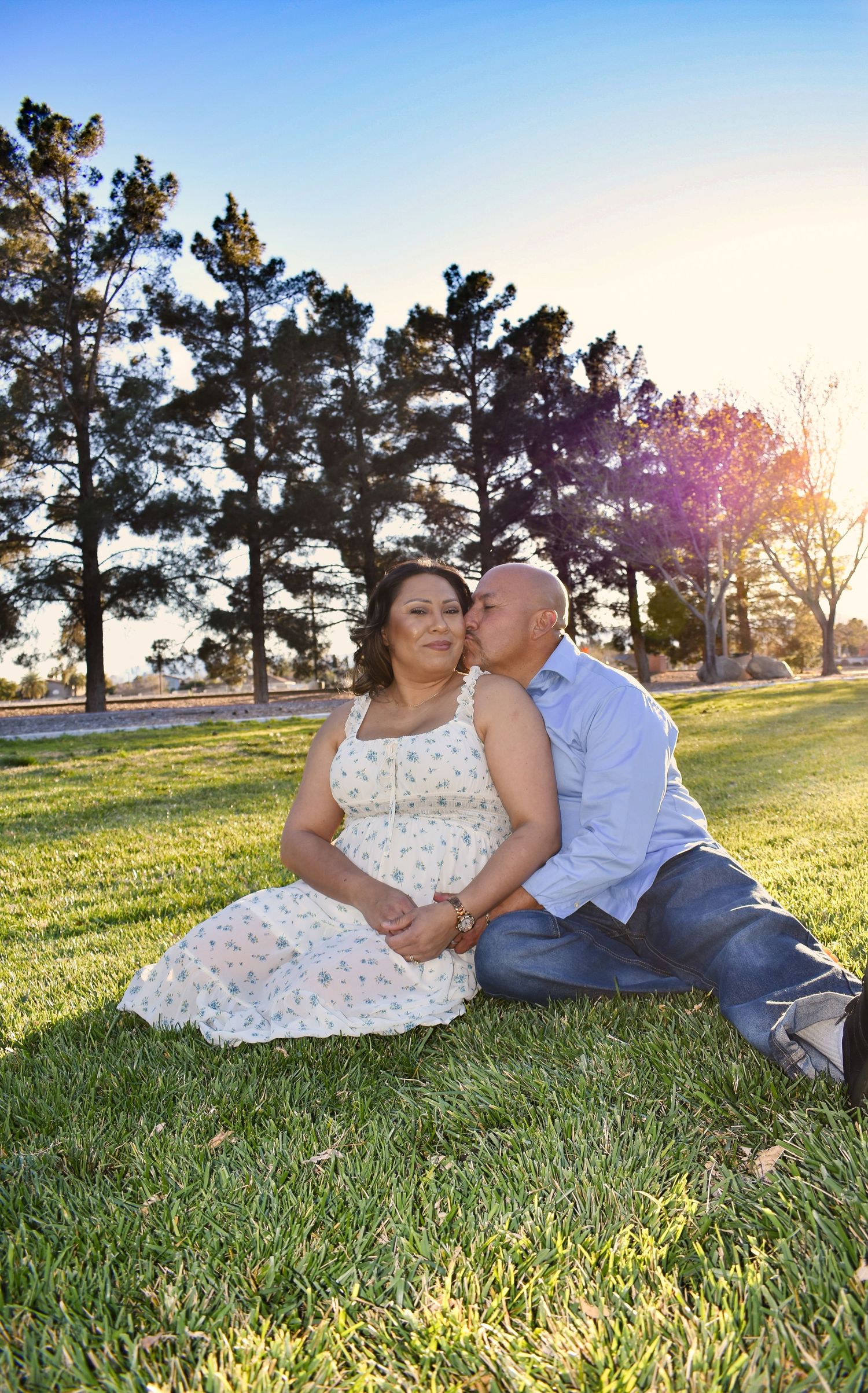 Couple sitting on green grass in a park, man kissing woman's cheek, trees and blue sky in background.