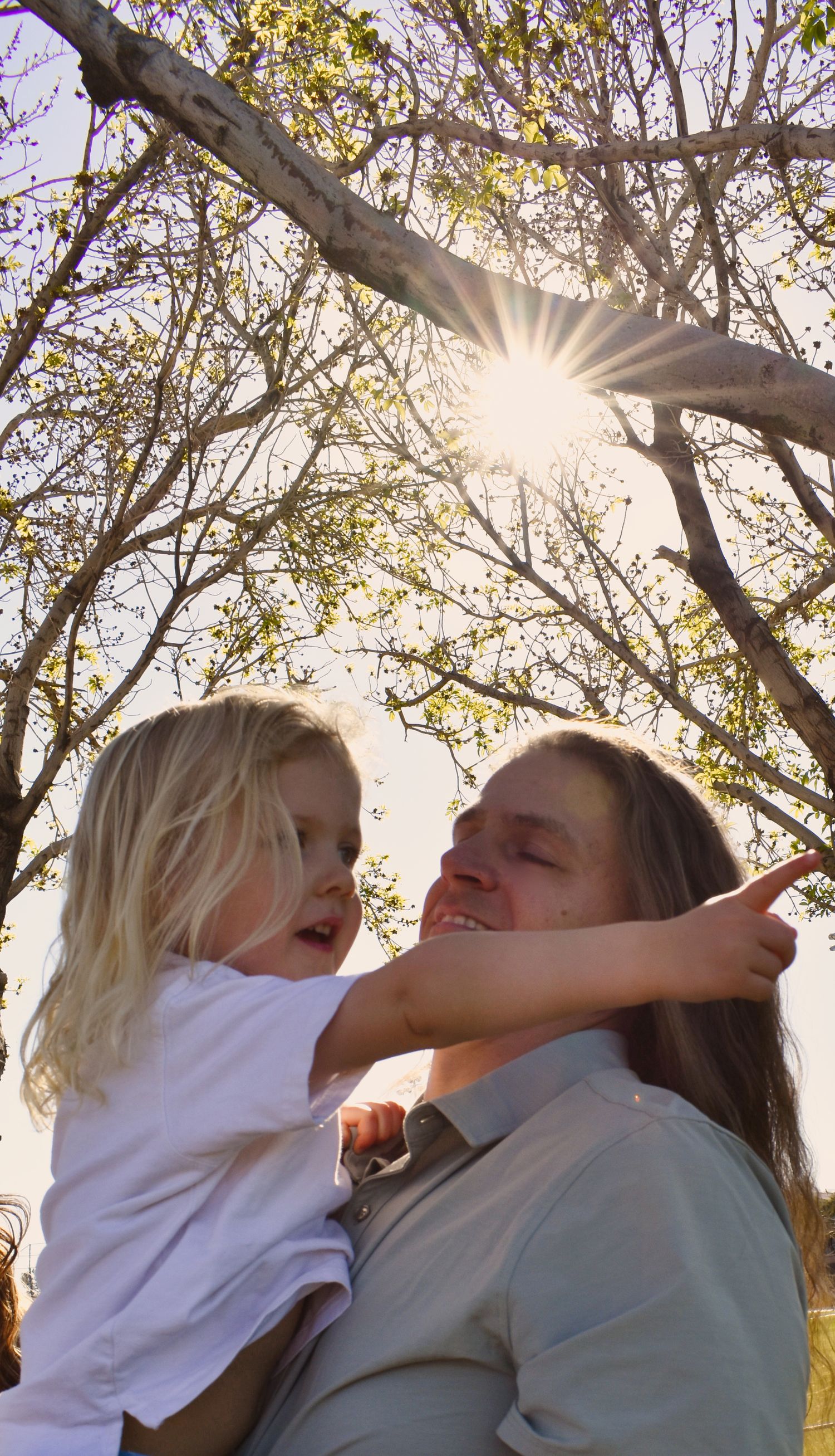 A couple embraces and laughs together under sunlit trees during an outdoor photoshoot.